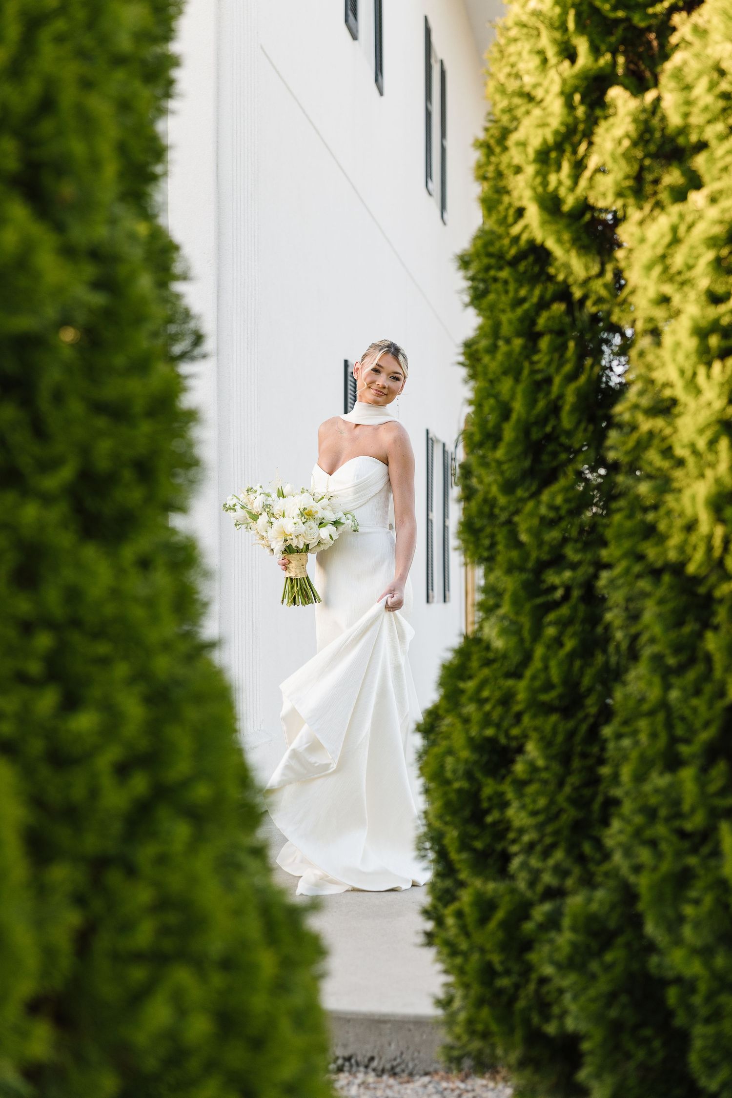 A figure in an elegant white wedding gown poses between lush green bushes along a white wall at an outdoor venue.