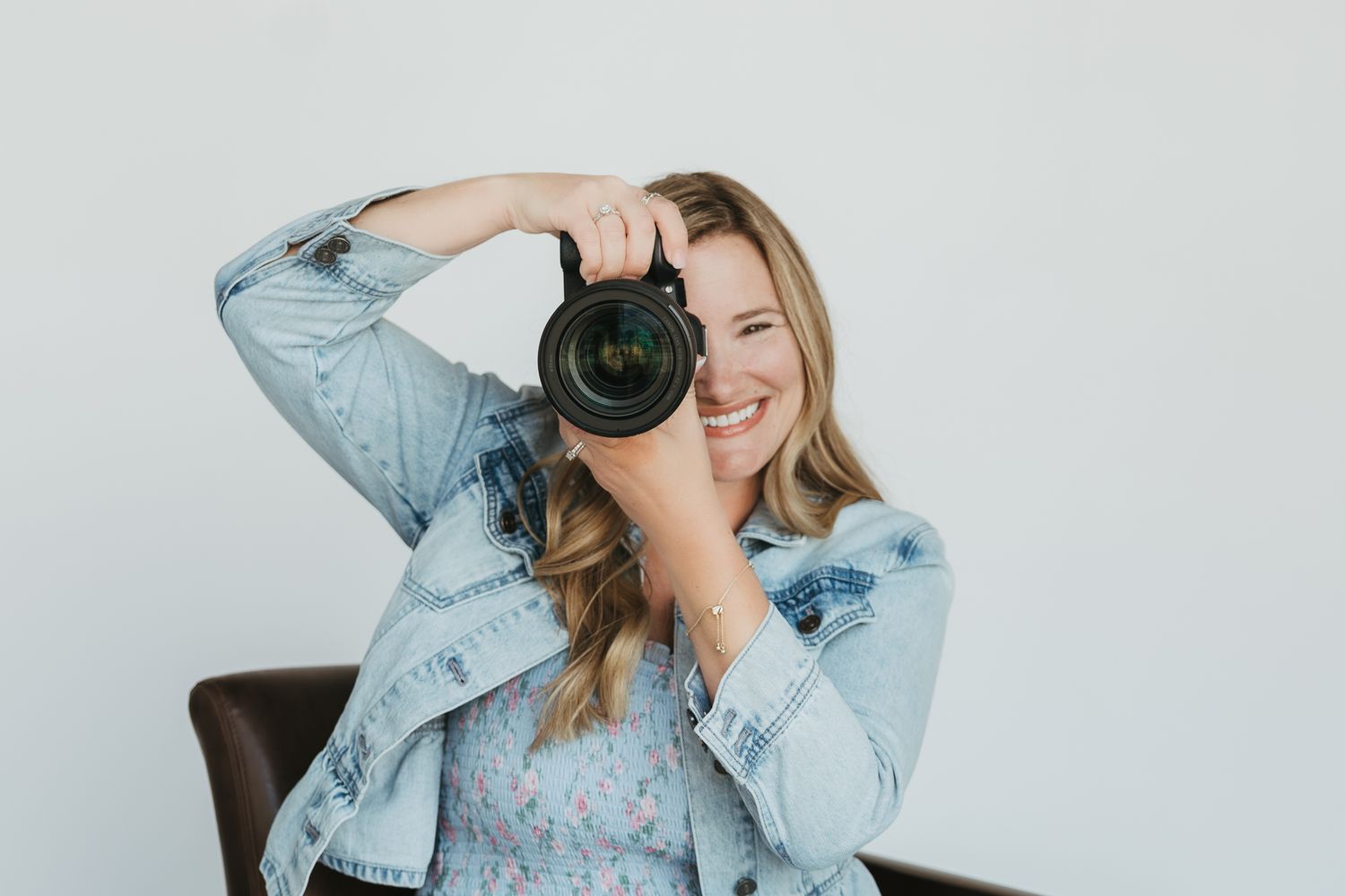 Photographer in light denim jacket smiling while taking photo with professional camera against white background.