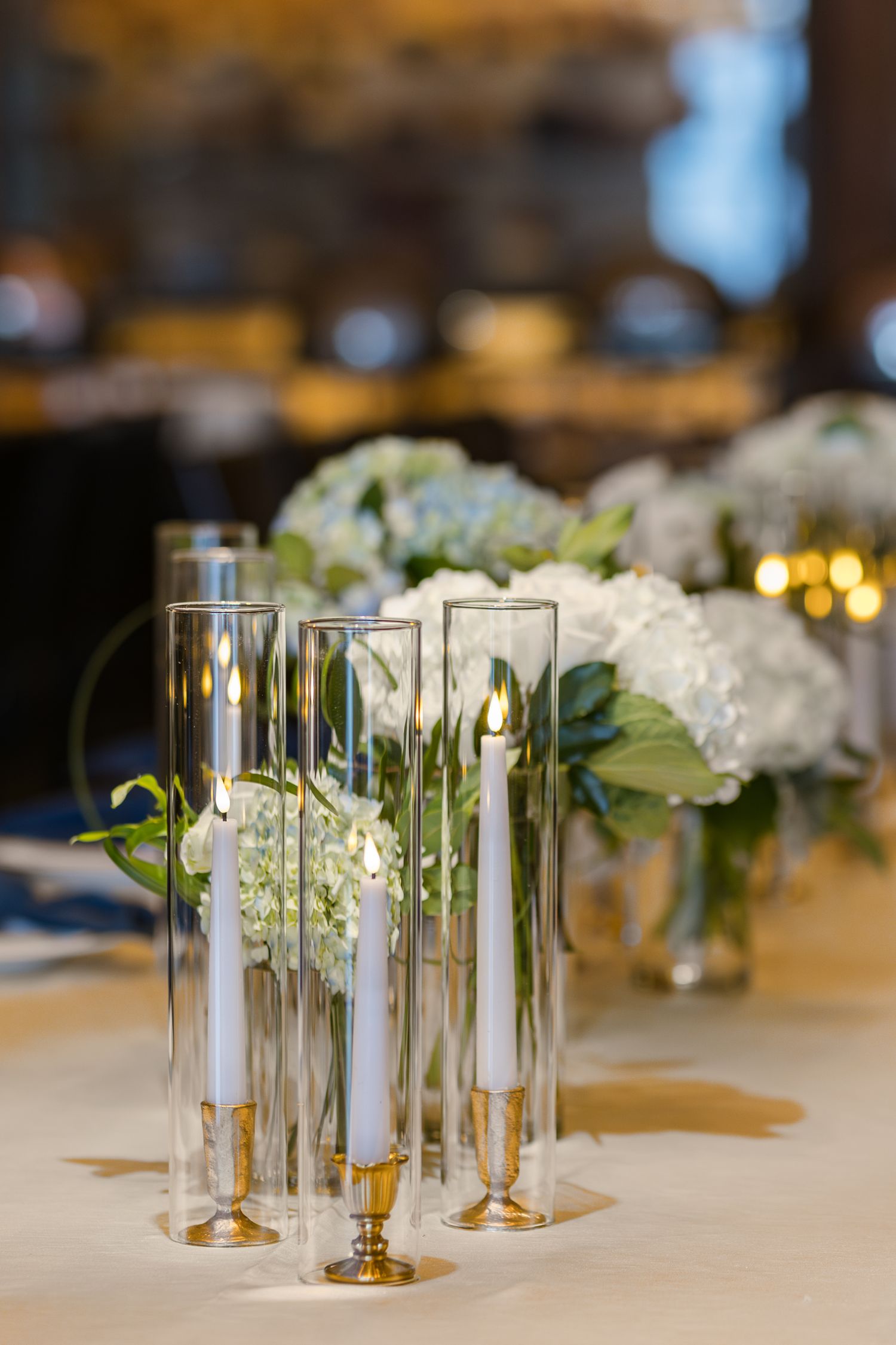 Elegant table setting with glass hurricane candle holders and white floral arrangements on a cream tablecloth.
