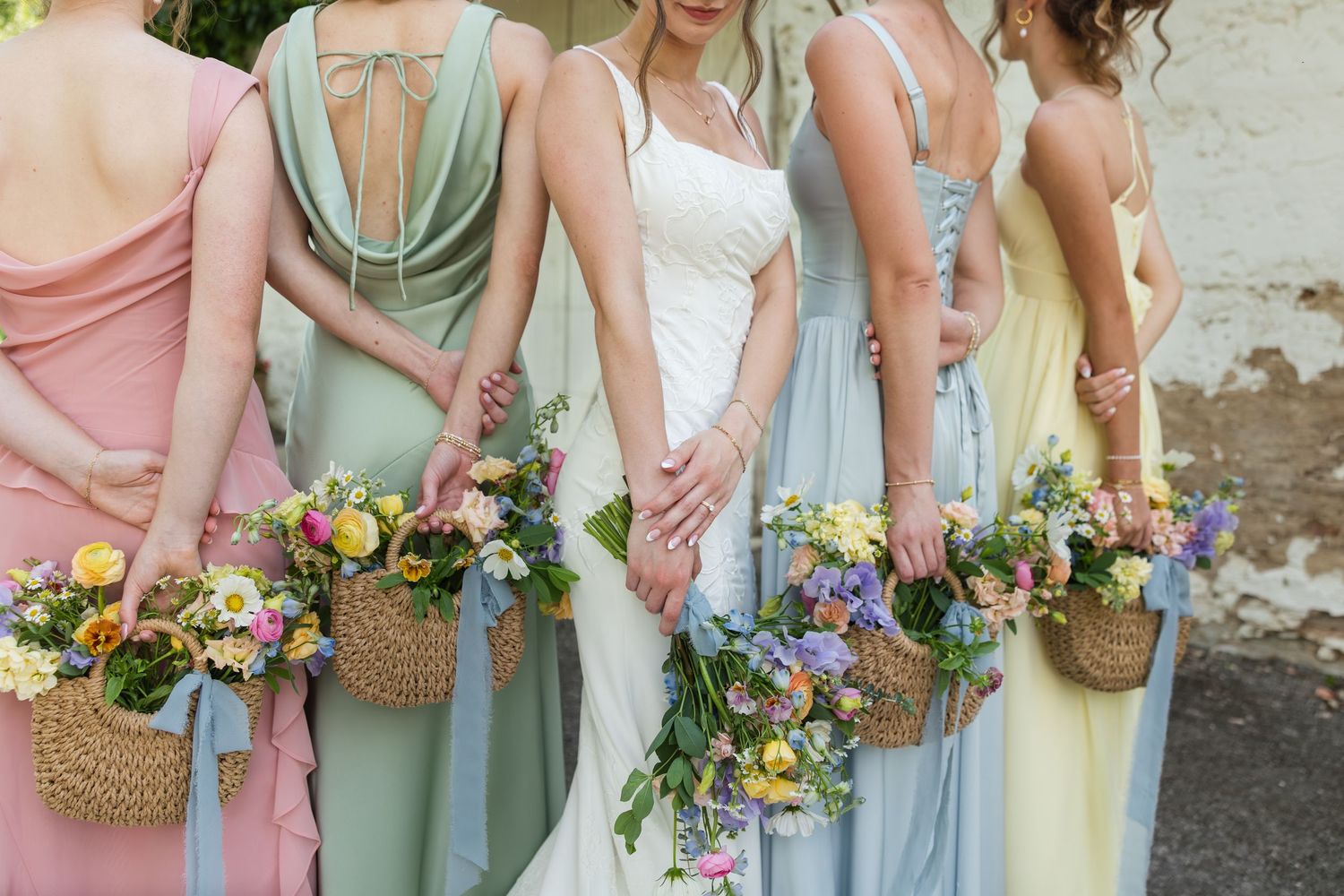 Bride in white gown with bridesmaids in pastel dresses holding wicker baskets of colorful wildflowers at a wedding.