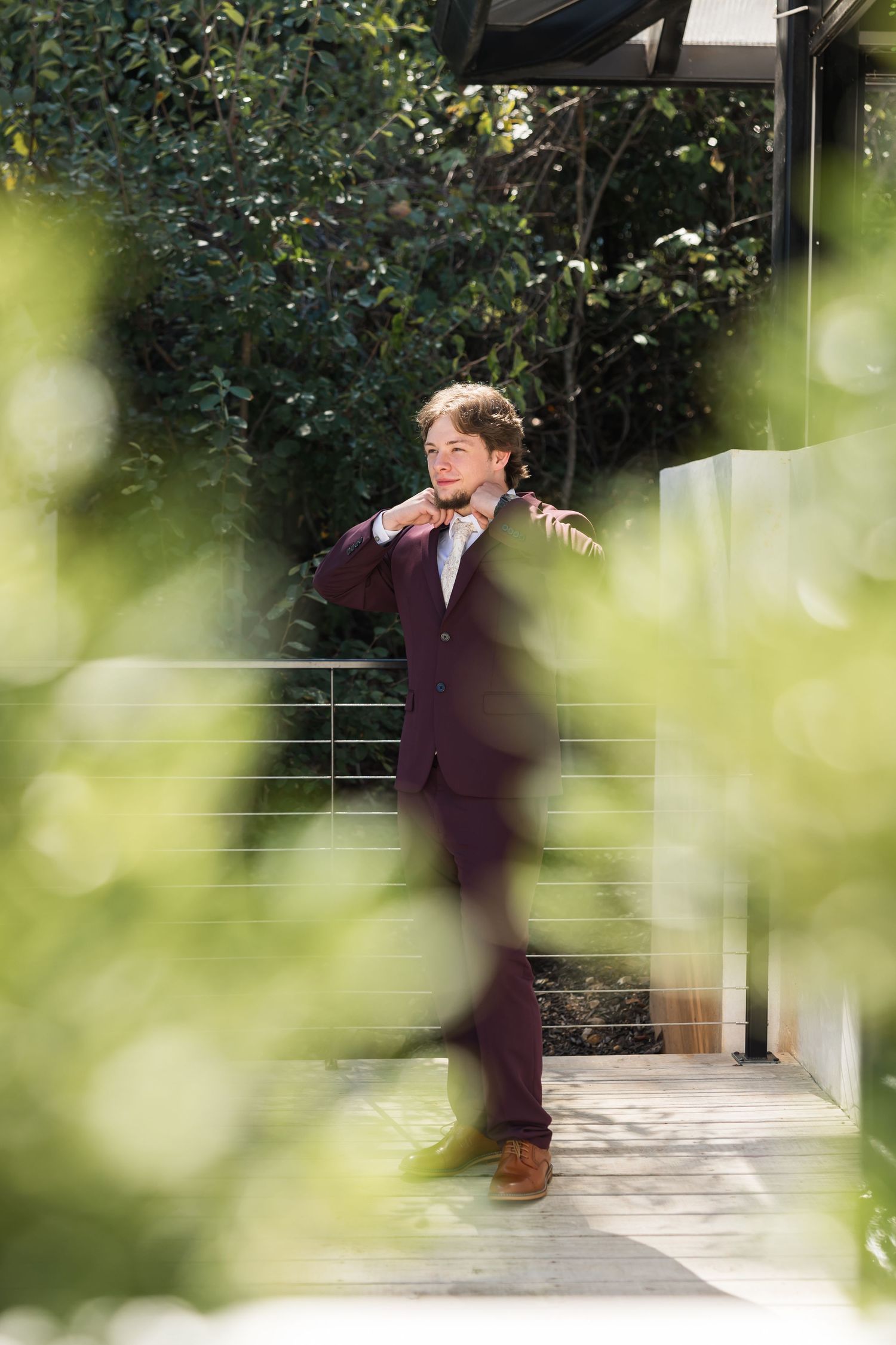 A groom in a maroon suit adjusts his tie outdoors, surrounded by lush greenery on a sunny day.