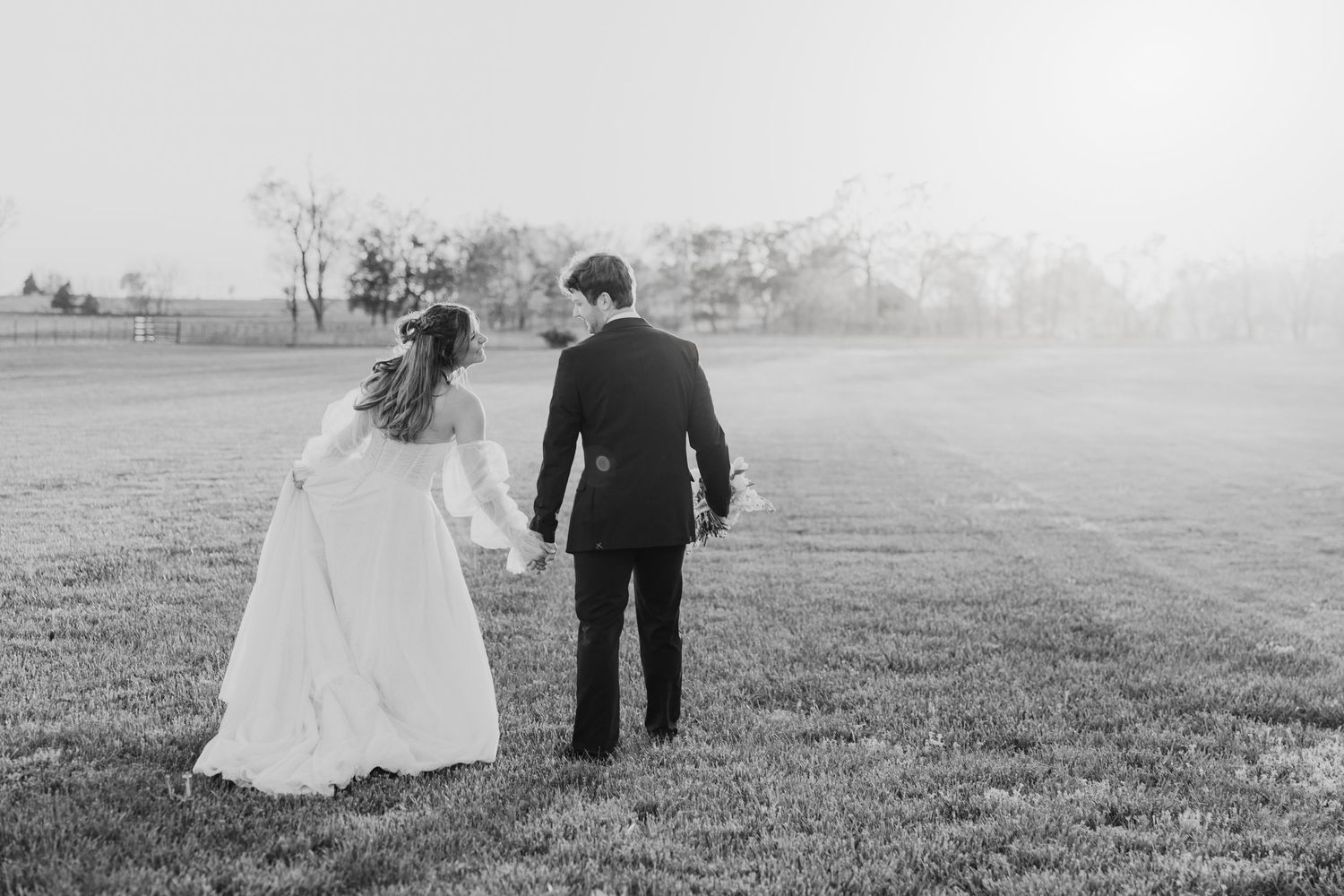 Black and white photo of a bride and groom holding hands walking through a misty field on their wedding day.