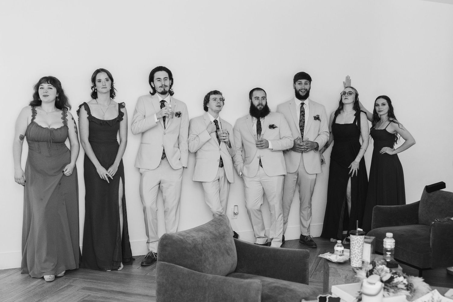 Black and white wedding party photo with groomsmen in suits and bridesmaids in gowns standing together indoors.