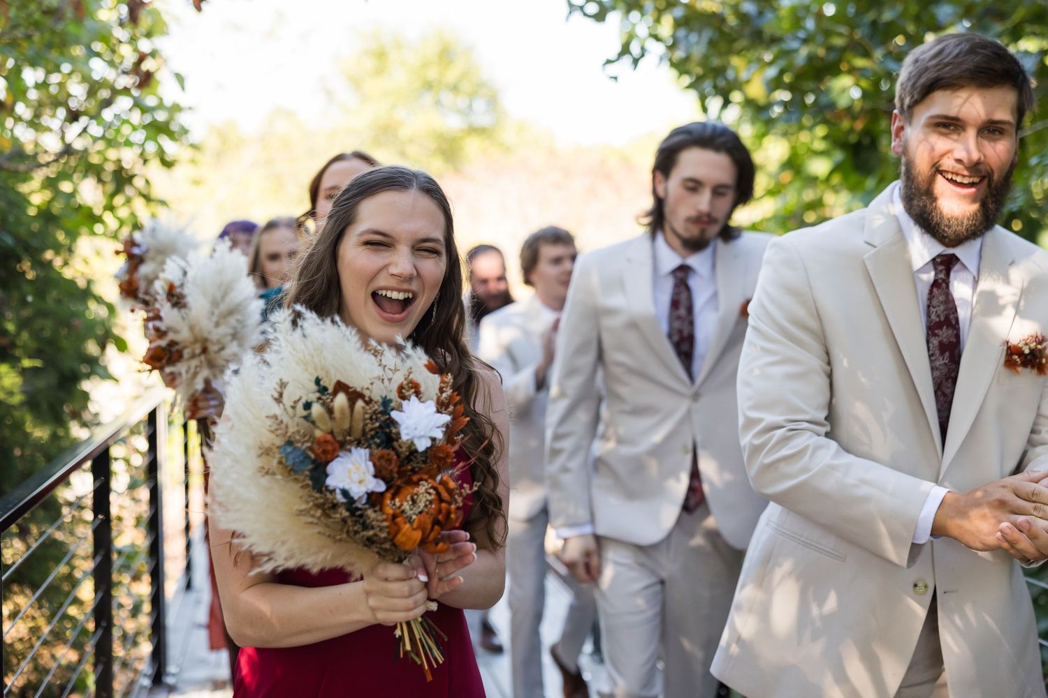 Smiling bridesmaid in red dress holds pampas bouquet beside bearded groomsman in tan suit outdoors.