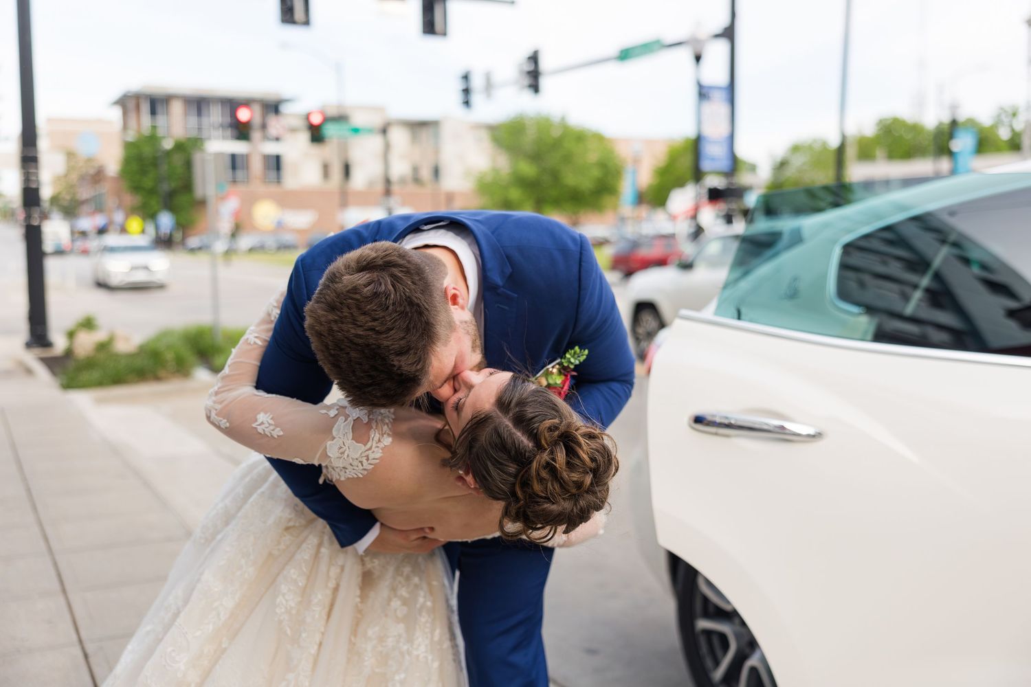 A groom dips and kisses his bride in a lace wedding gown on a city sidewalk beside a white car.