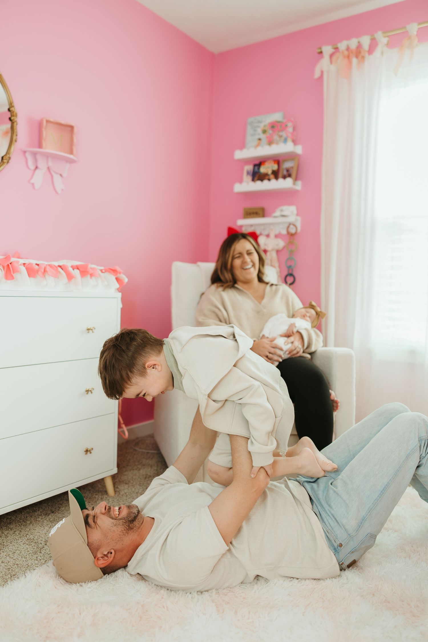 A family shares a playful moment together on the floor of a pink nursery decorated with white furniture and shelving.