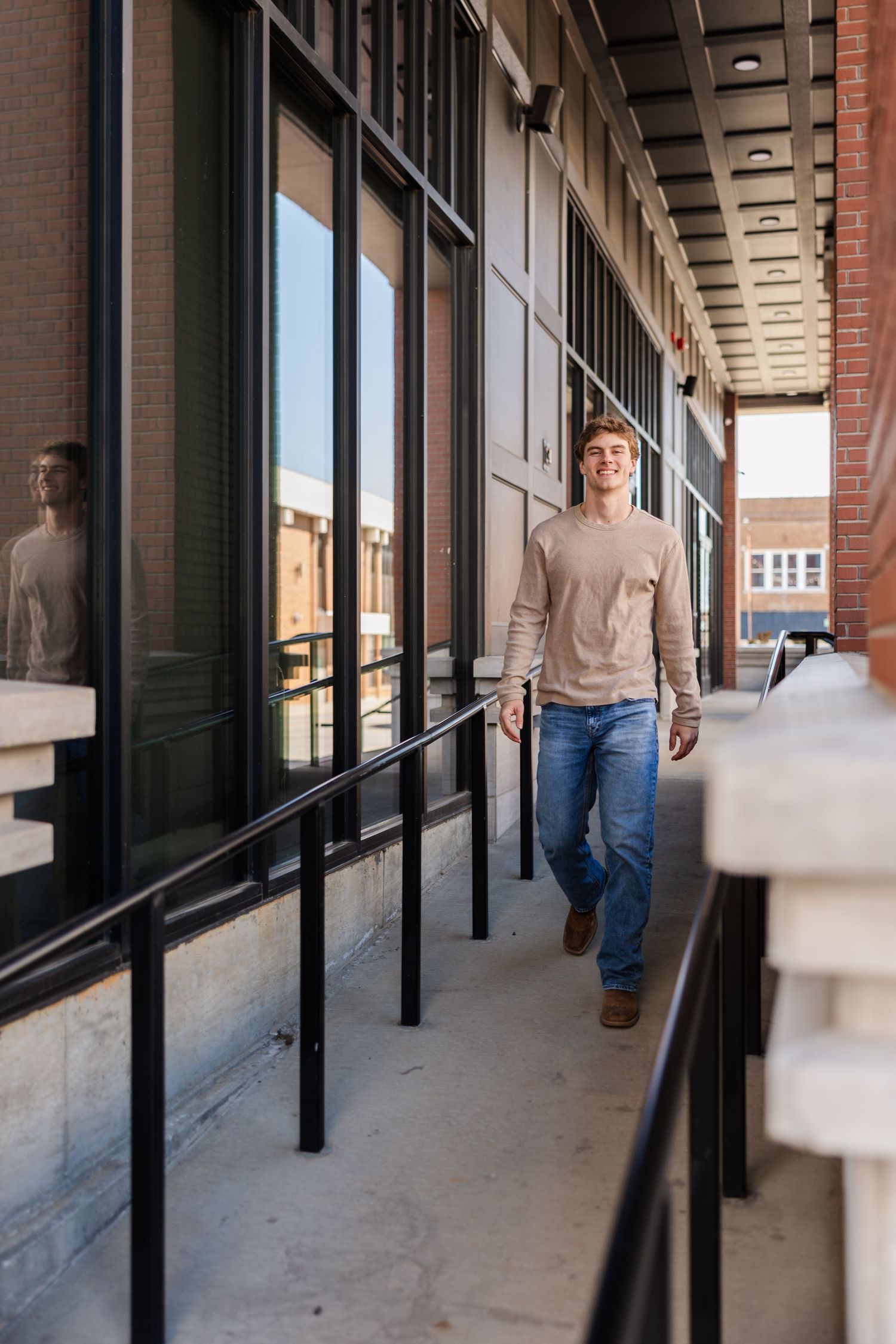 Person in beige sweater walking along modern building corridor with reflective windows.