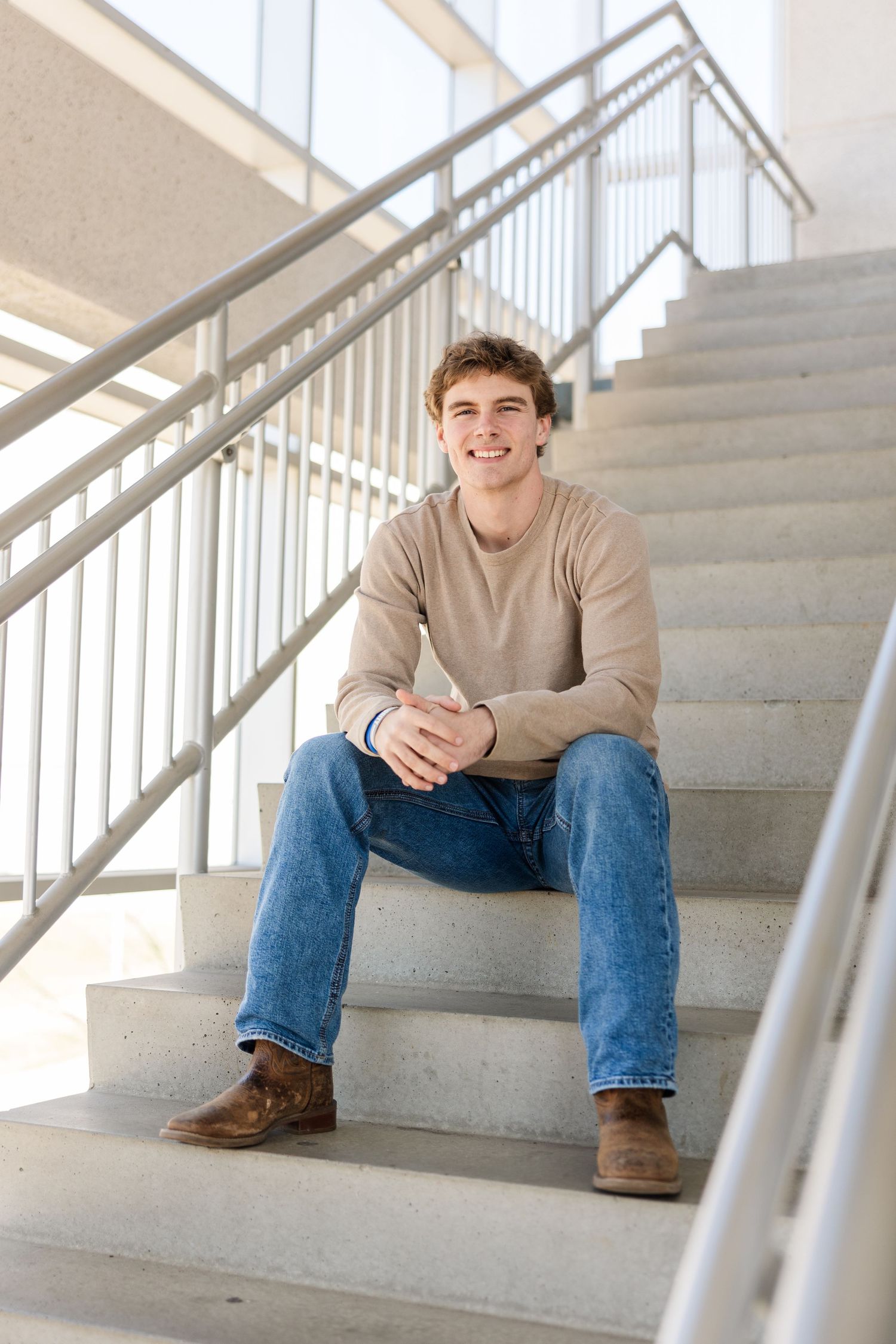 Person wearing beige sweater and jeans sits casually on modern concrete stairs with metal railing.