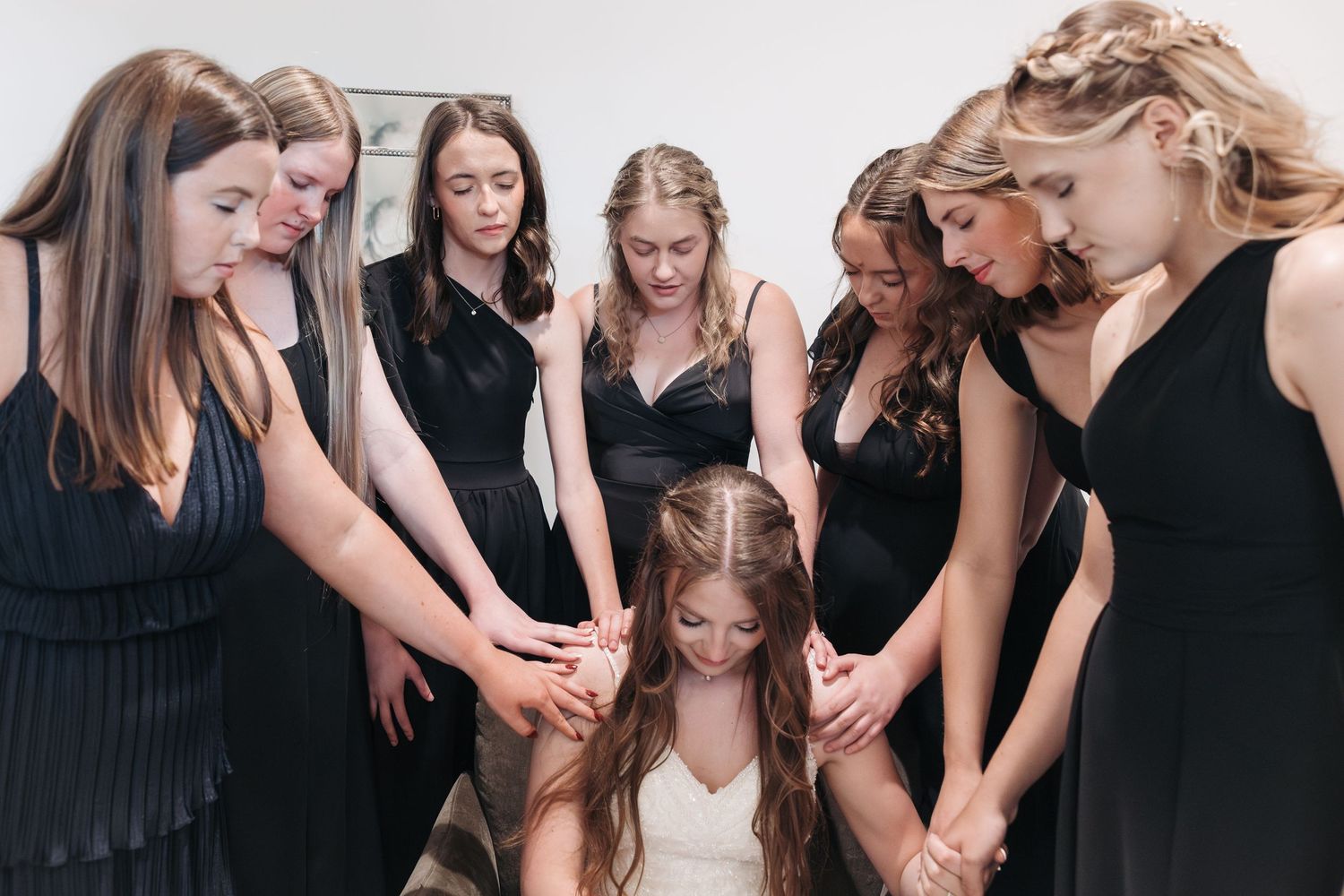 Wedding party in black dresses gathers around the bride in white for an emotional prayer circle moment.