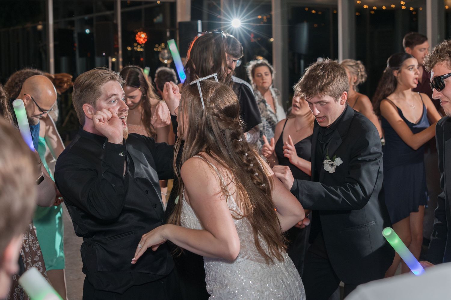 Wedding guests dance energetically with light-up sticks at an indoor reception.
