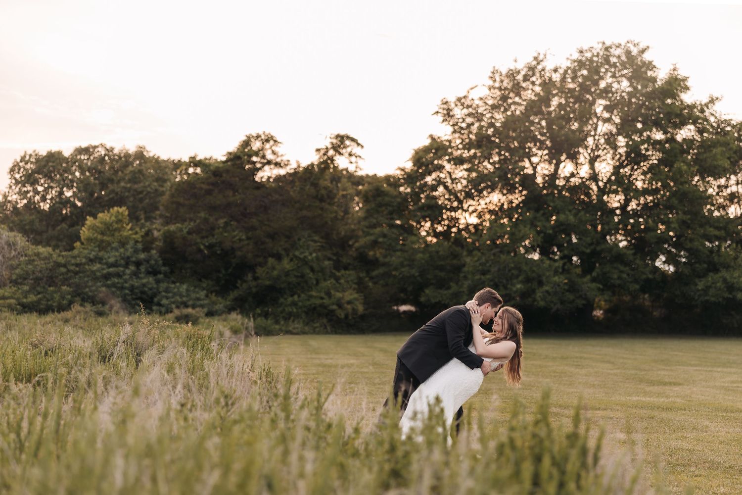 A romantic dip pose between newlyweds in a grassy field at sunset.