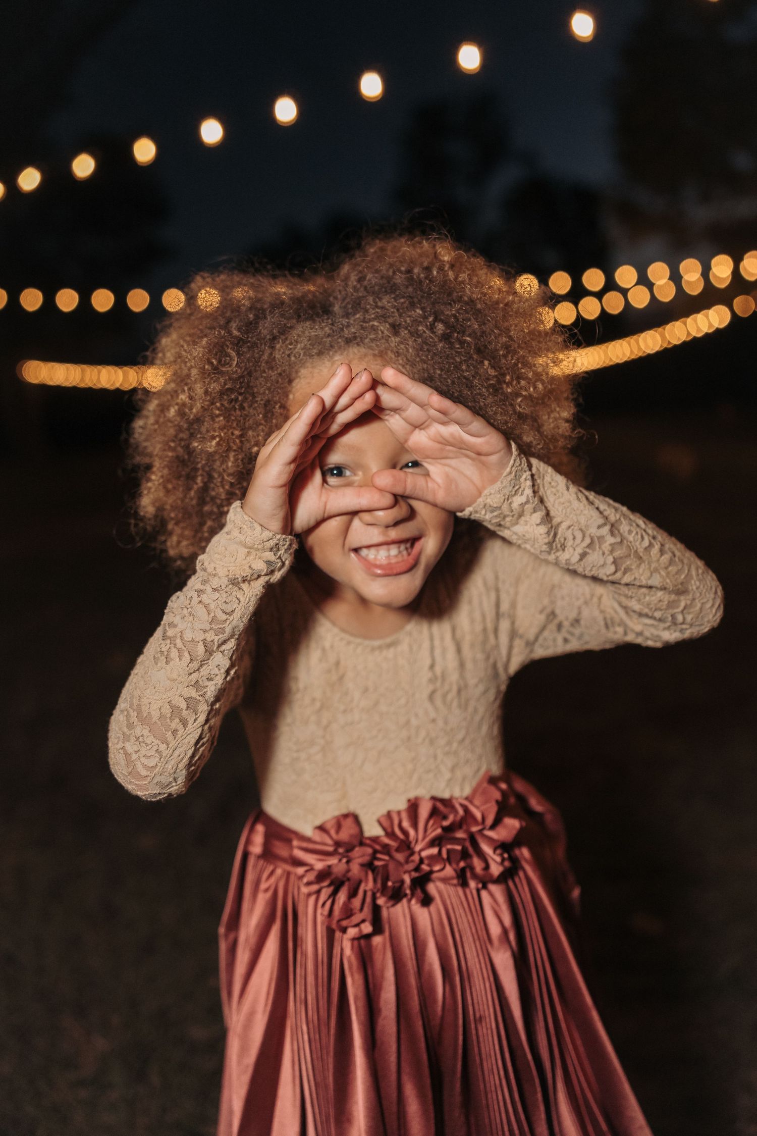 Joyful nighttime portrait under string lights wearing cream sweater and mauve skirt.