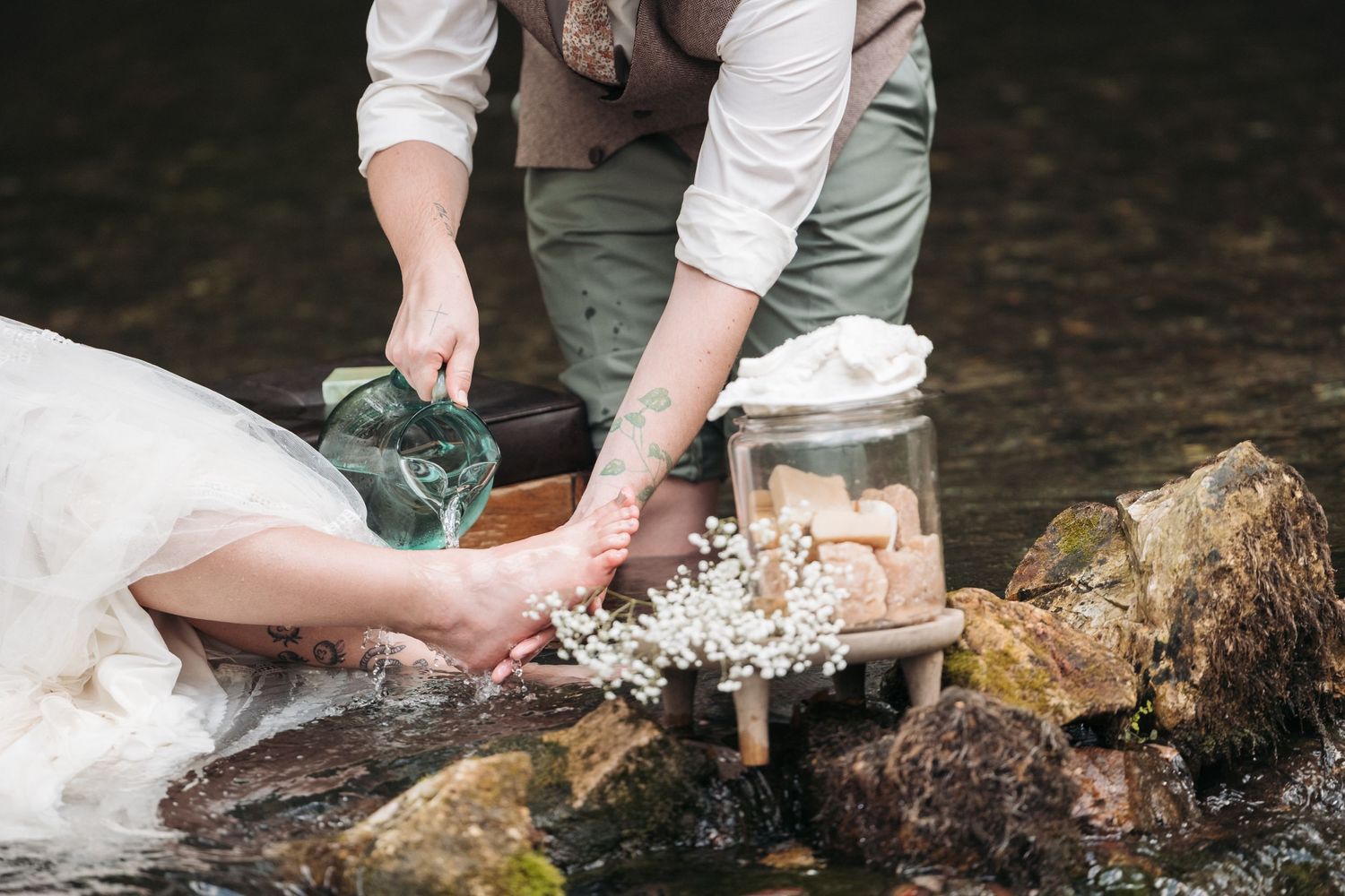 Someone in a wedding dress pours water from a glass vessel during an outdoor ceremony.