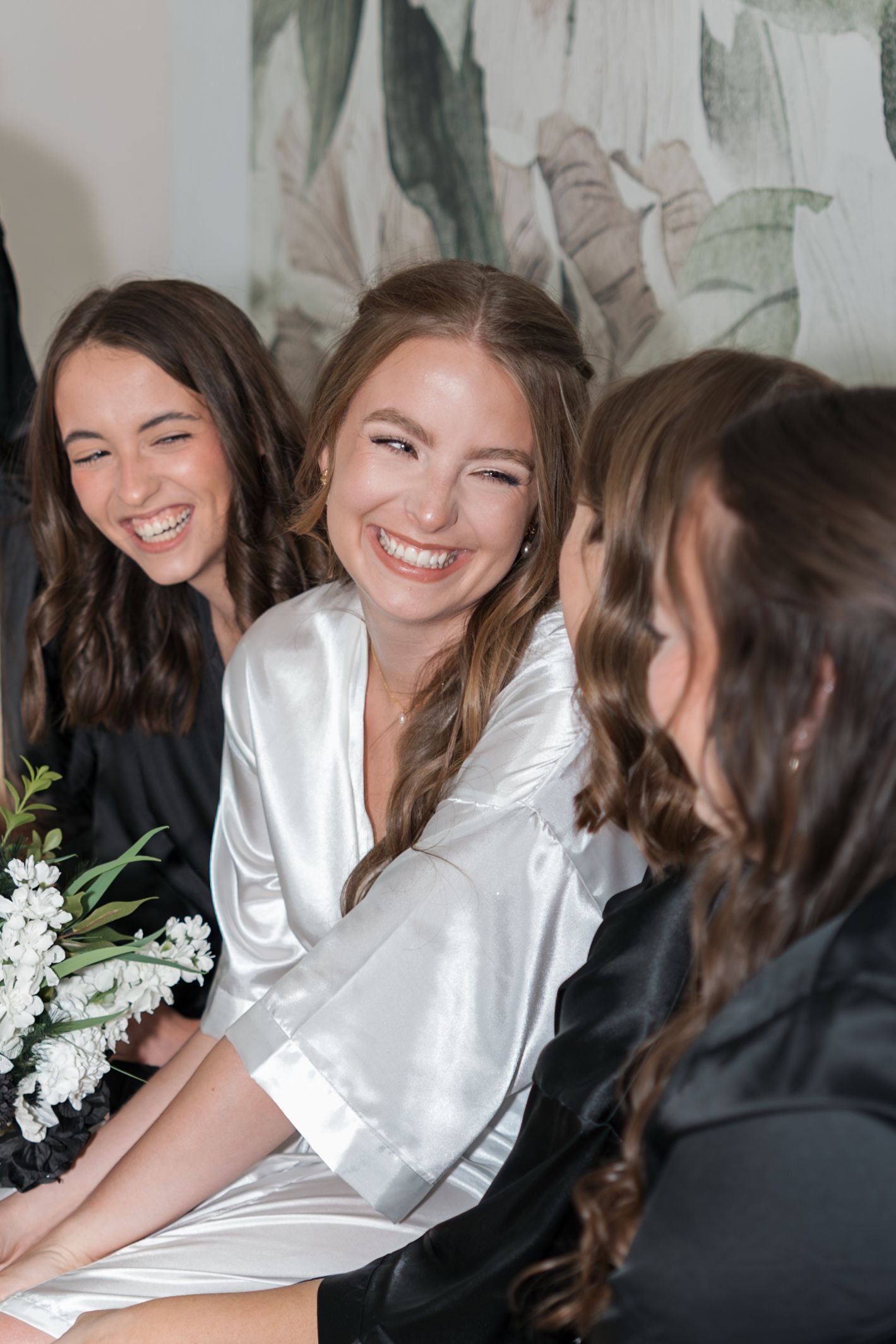 Bridesmaids sharing joyful laughter while getting ready in white silk robes and black dresses.