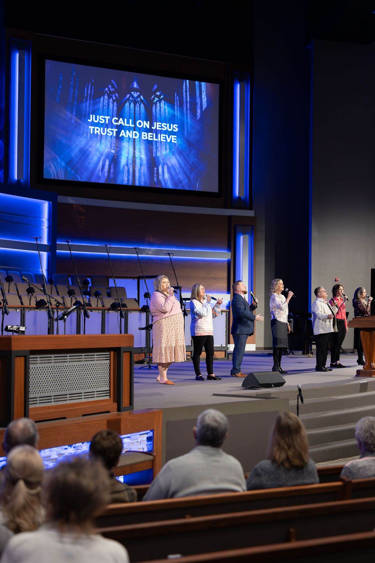 Church worship service with choir members singing and projection screen displaying lyrics in blue lighting.