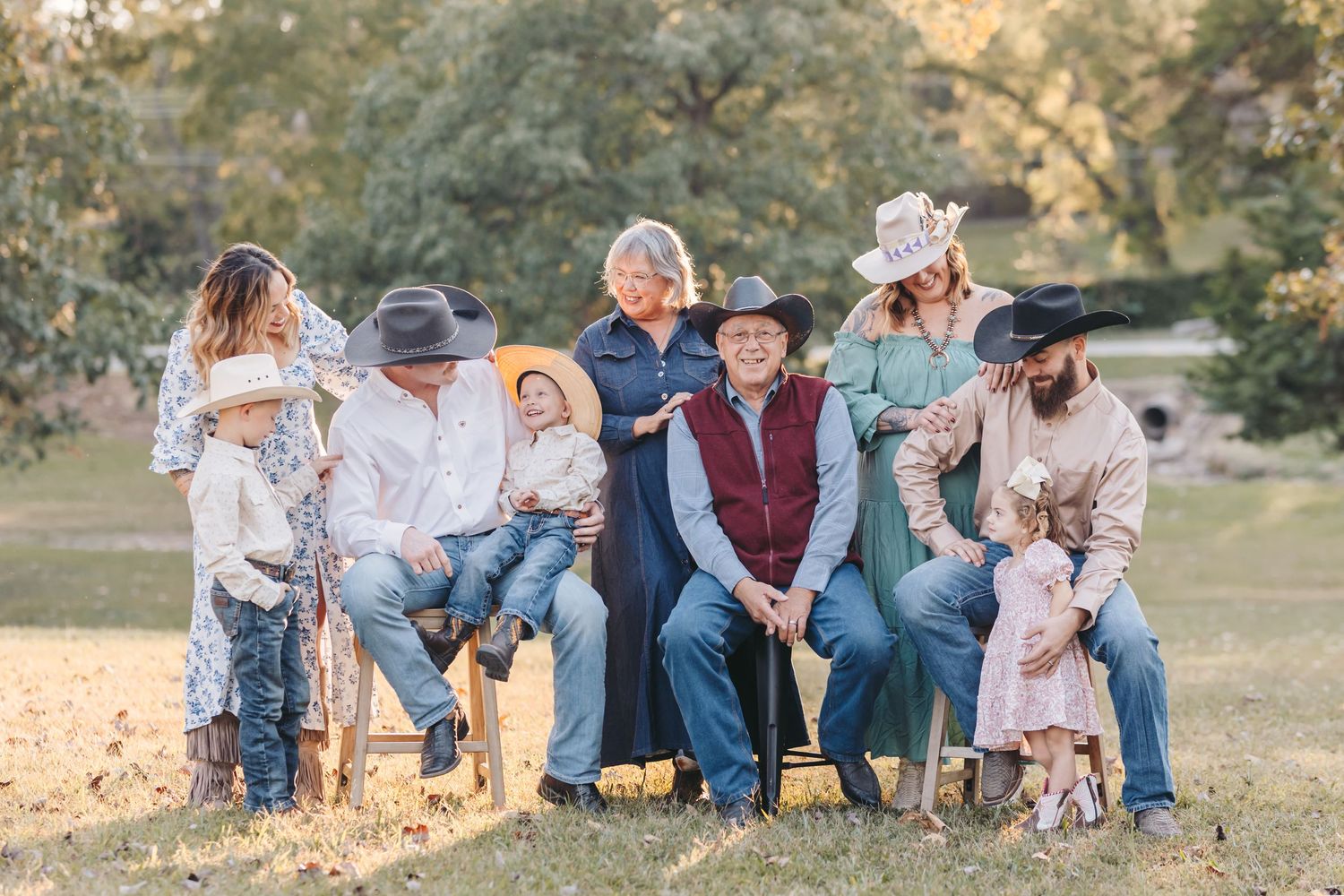 Extended family members sit together on wooden stools in a park during sunset.