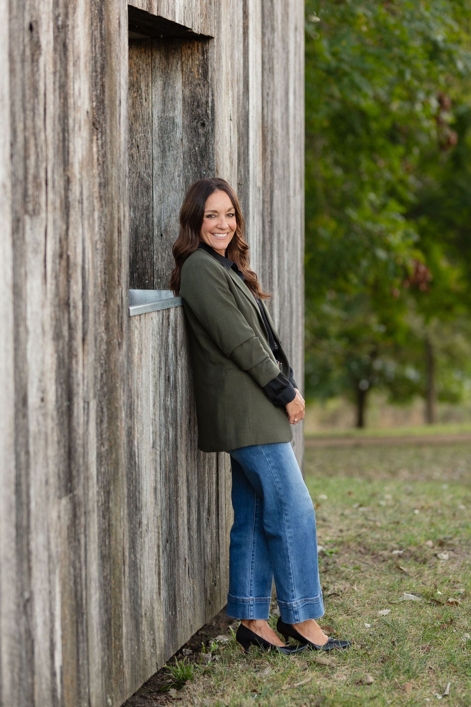 Person in blazer and jeans poses against rustic wooden wall outdoors.