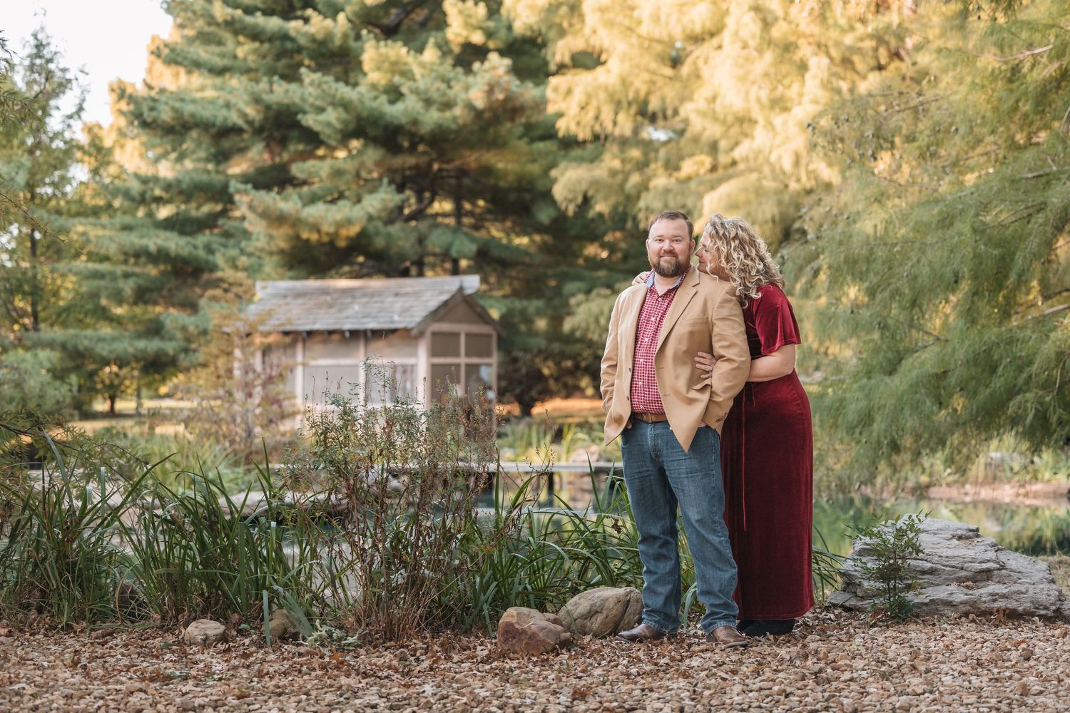 Couple standing near a rustic cabin surrounded by evergreen trees in warm autumn lighting.