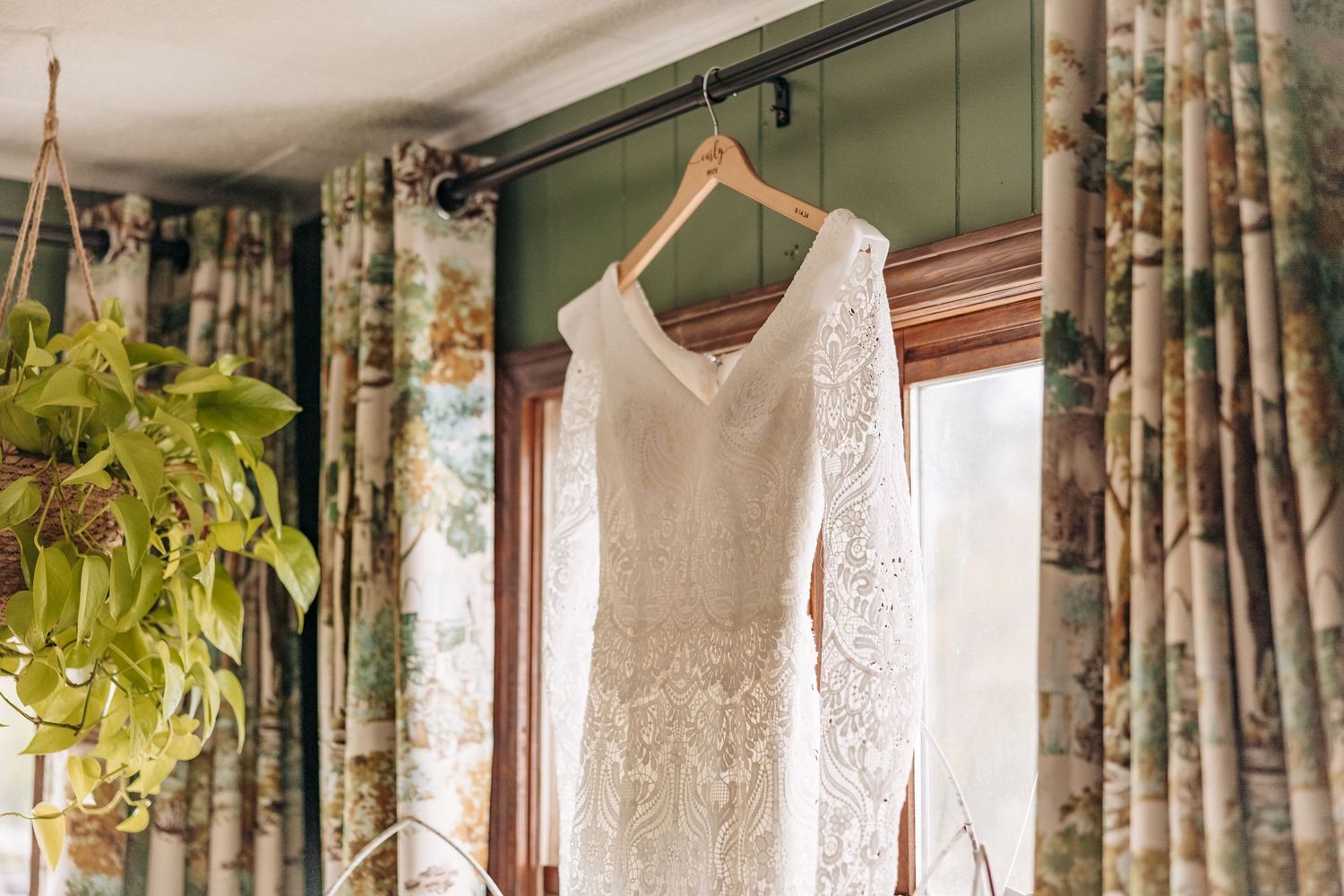 A delicate lace wedding dress with long sleeves hangs from a wooden hanger against floral curtains near a houseplant.