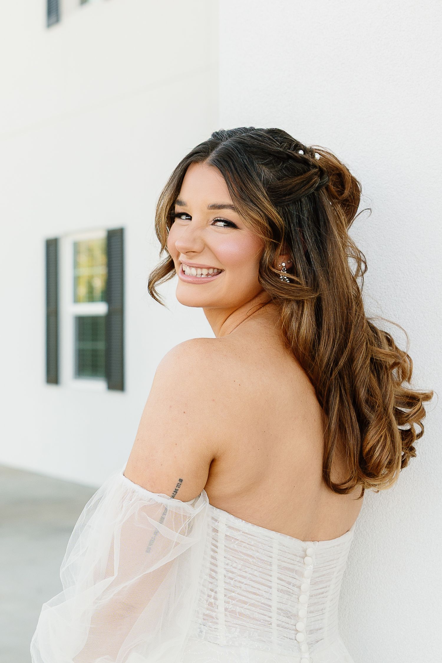 Bride in elegant off-shoulder white wedding dress with corset back and flowing curls smiles during outdoor photoshoot.