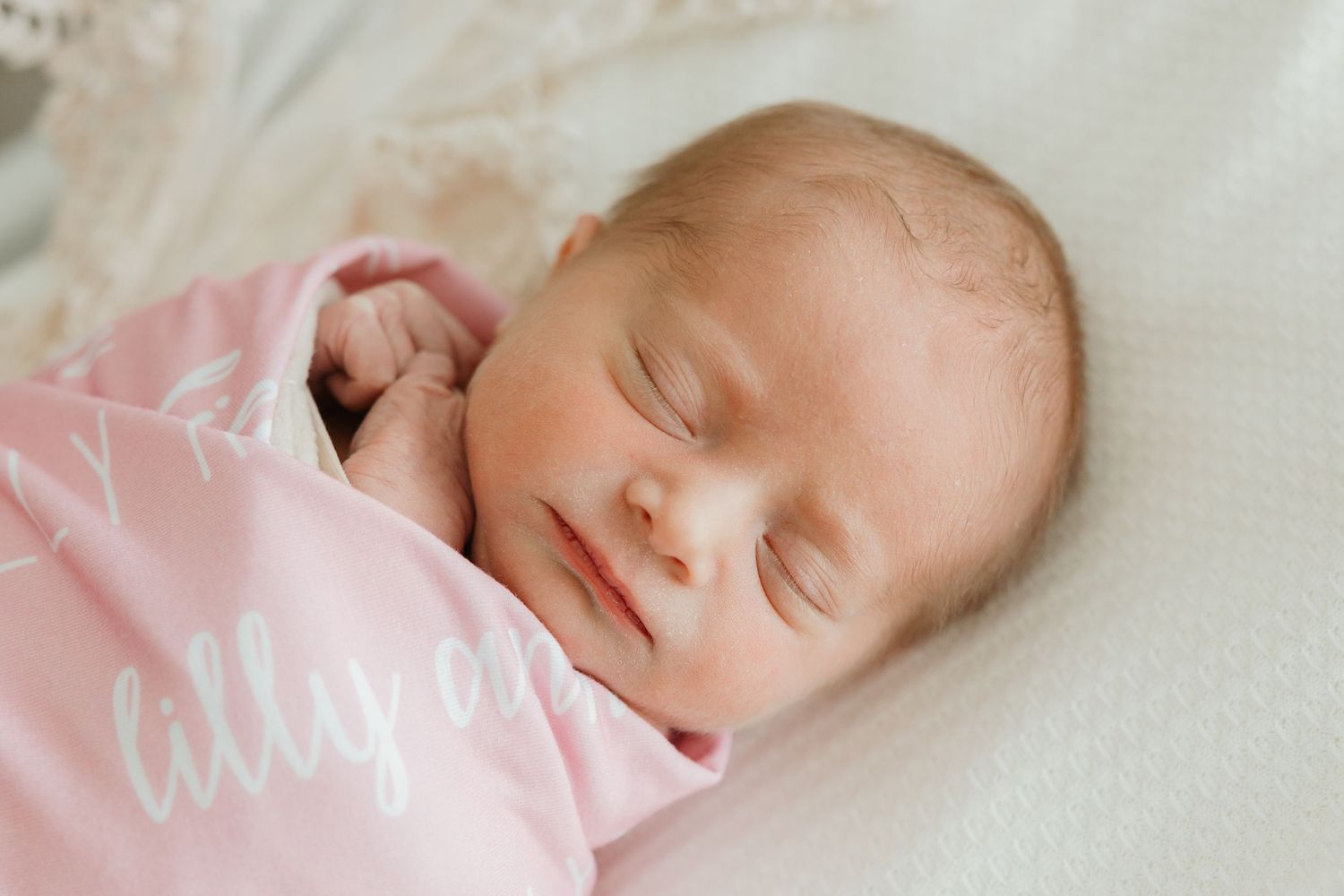 A sleeping newborn baby wearing pink swaddle blanket rests peacefully on white bedding.