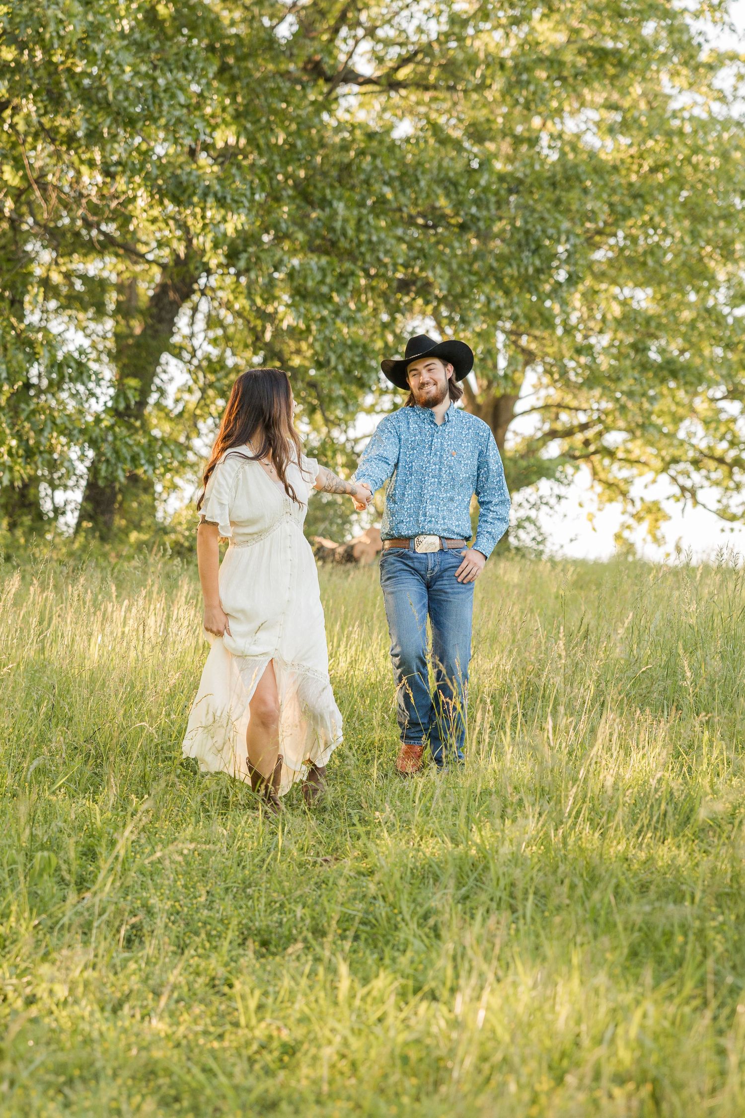 A couple walks hand in hand through a sunlit meadow with tall grasses and trees during golden hour.