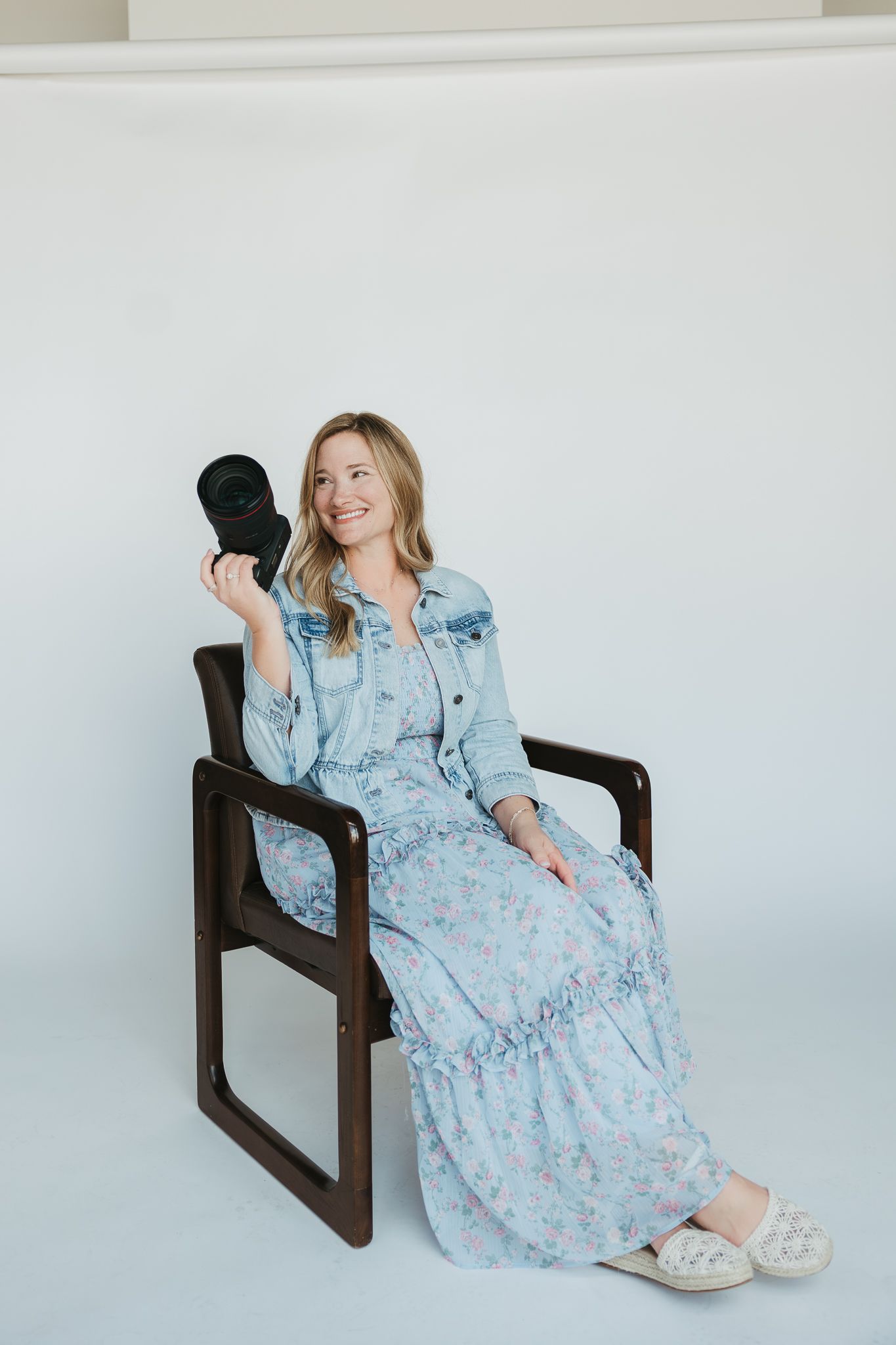 Photographer in blue floral maxi dress and denim jacket holds camera lens while sitting in modern chair.