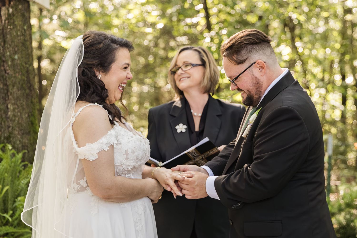 Bride and groom exchange rings during outdoor forest wedding ceremony with smiling officiant holding book.