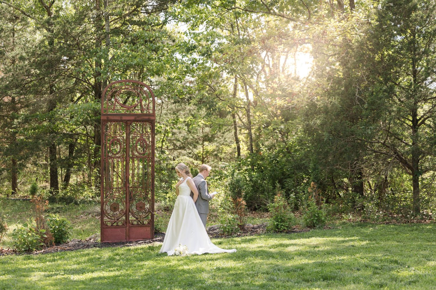 Bride and groom share a romantic kiss in a lush garden beside an ornate iron gate, with sunlight filtering through trees.
