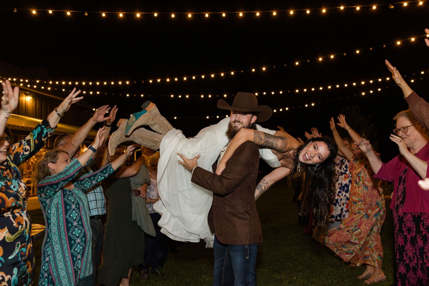 Groom lifts bride surrounded by cheering guests under string lights at outdoor wedding reception at night.