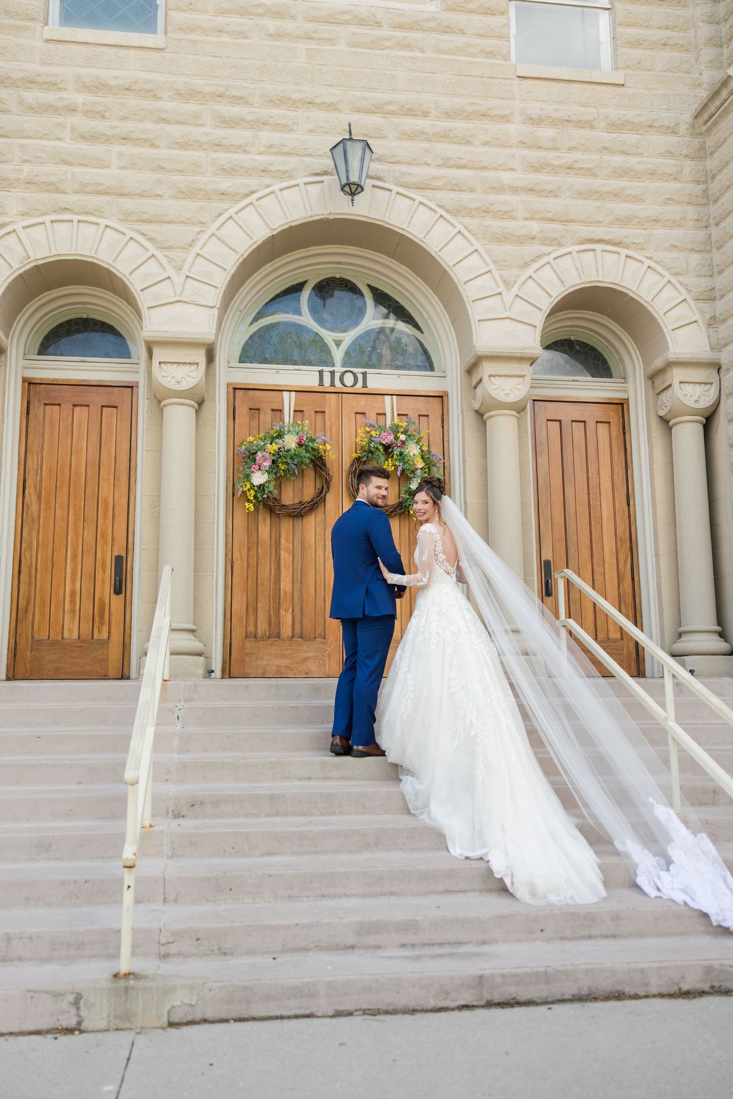 Bride and groom pose on church steps, her veil flowing, in front of arched wooden doors at 1101.