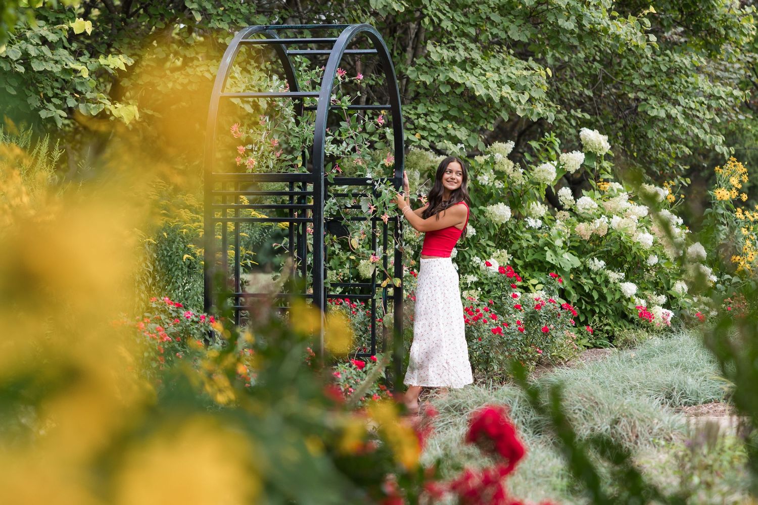 Woman in red top and white skirt standing in a lush garden with colorful roses and a black metal arch.