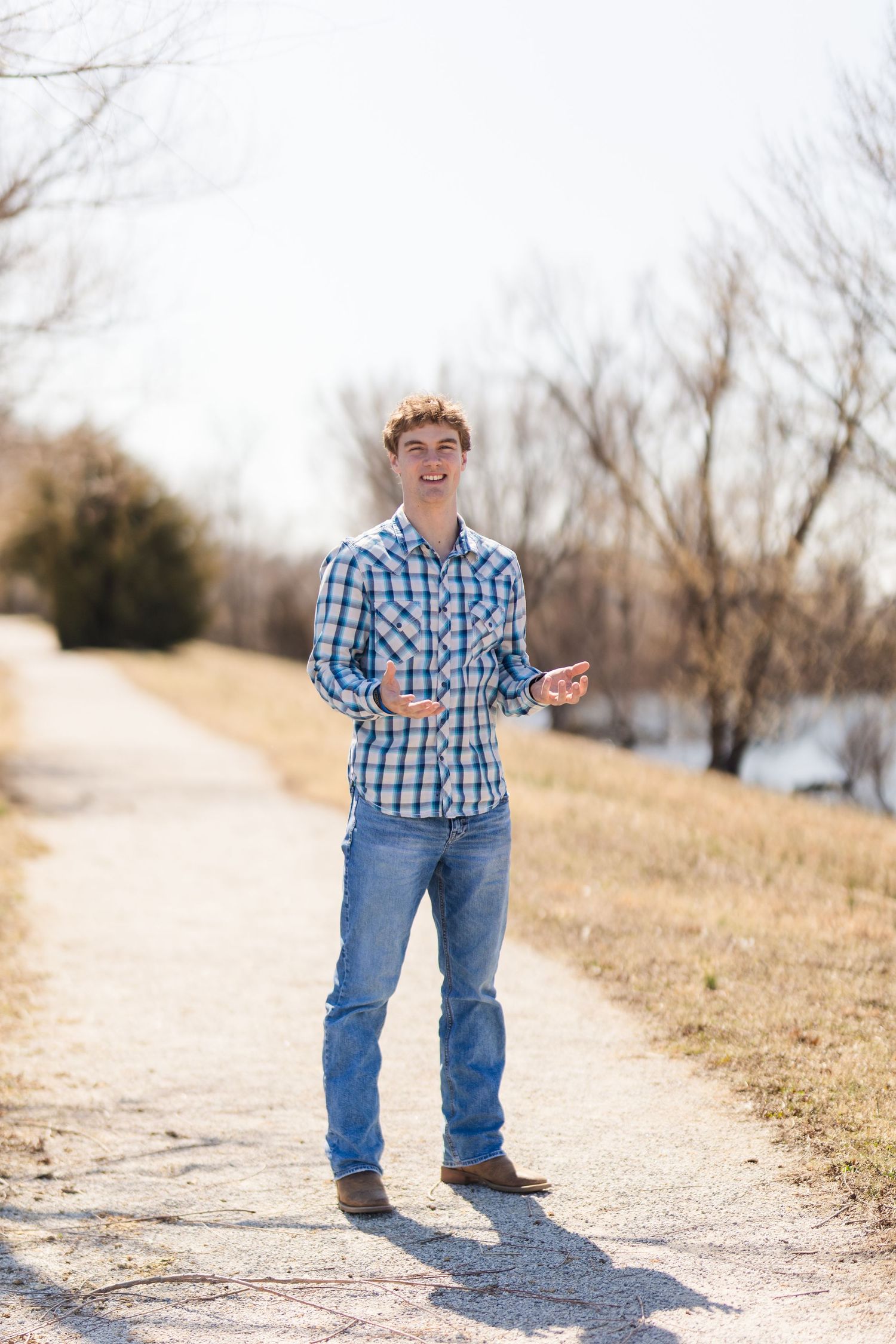 Person in blue plaid shirt and jeans standing on path with winter trees and river in background.