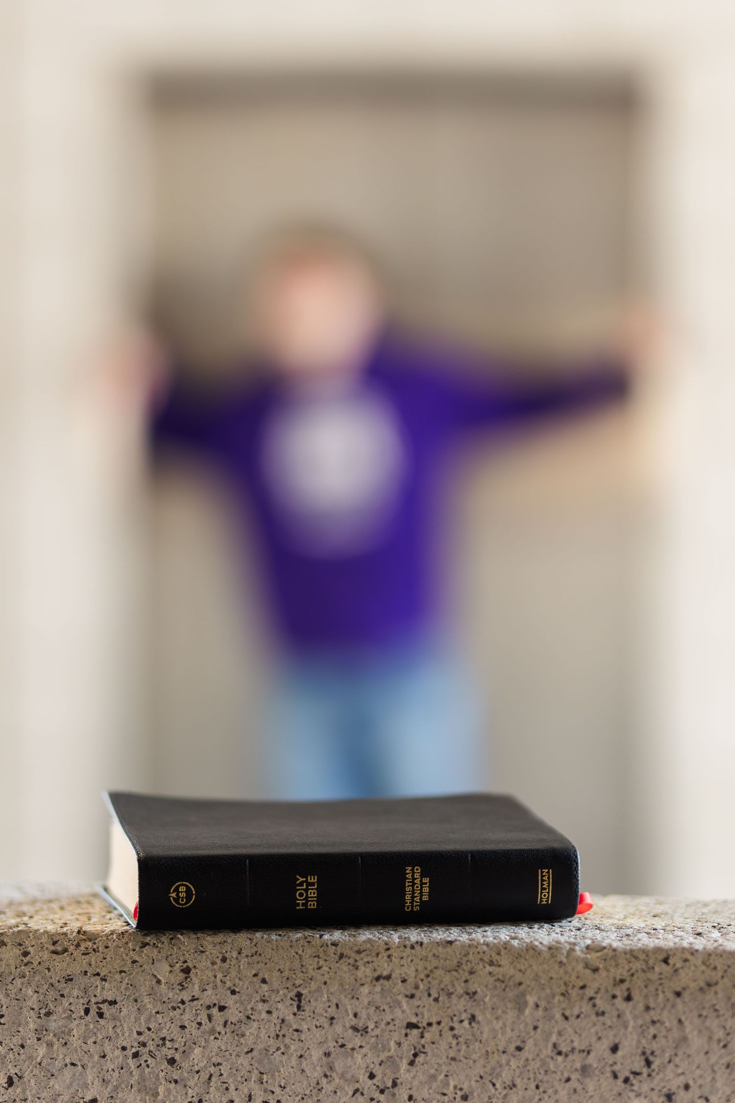 A black Bible sits in focus on a concrete ledge with a blurred purple-clad figure in the background.
