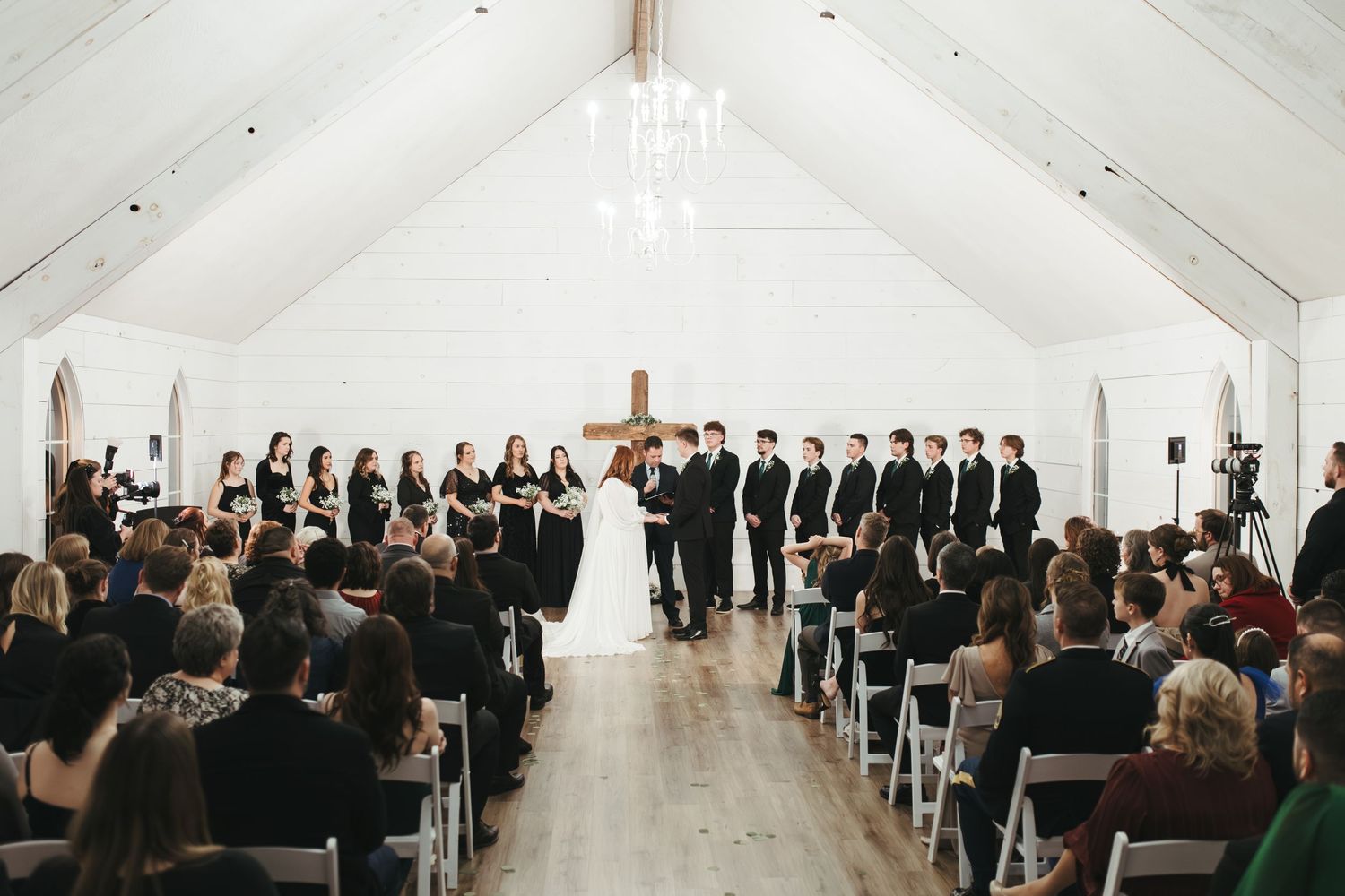 A wide shot of a wedding ceremony in a bright white chapel with guests seated and wedding party at the altar.