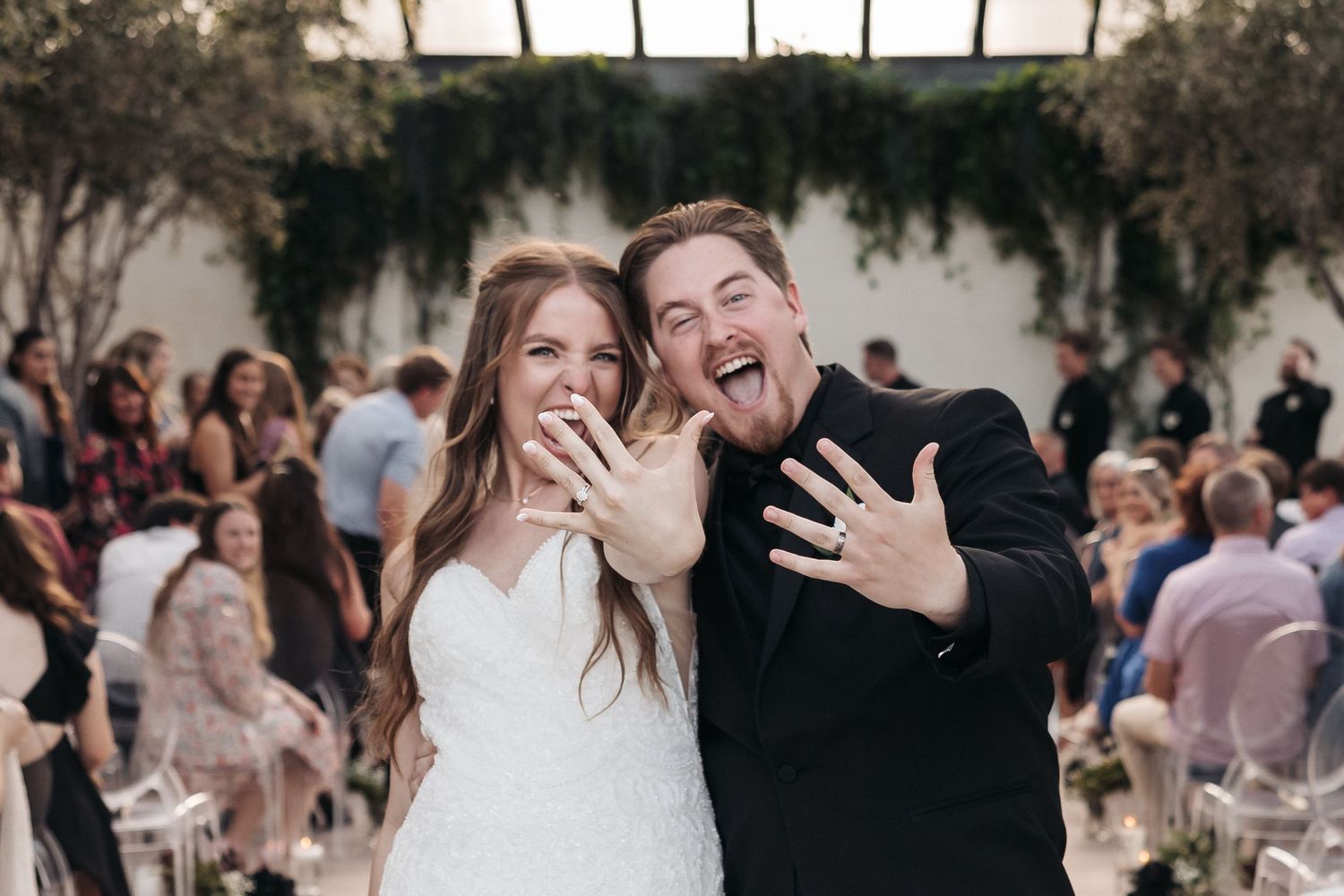 Newlyweds excitedly showing off their wedding rings at an outdoor ceremony with guests in the background.