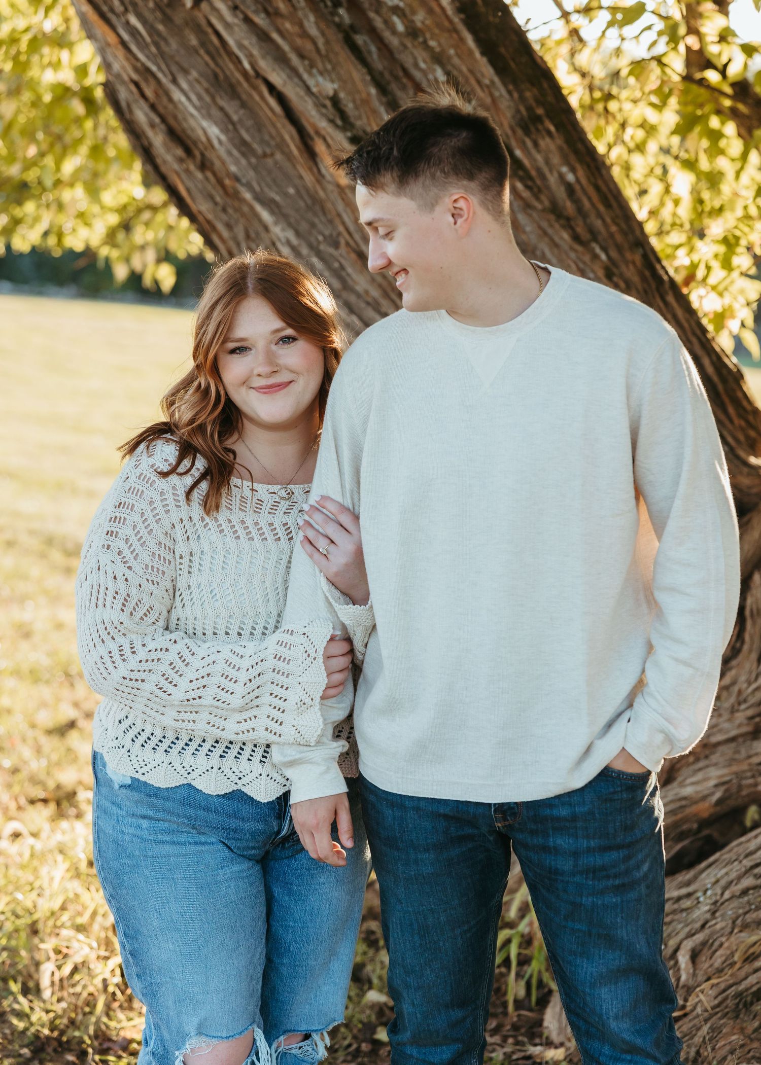 A couple shares a joyful moment while standing by a large tree in golden sunlight.