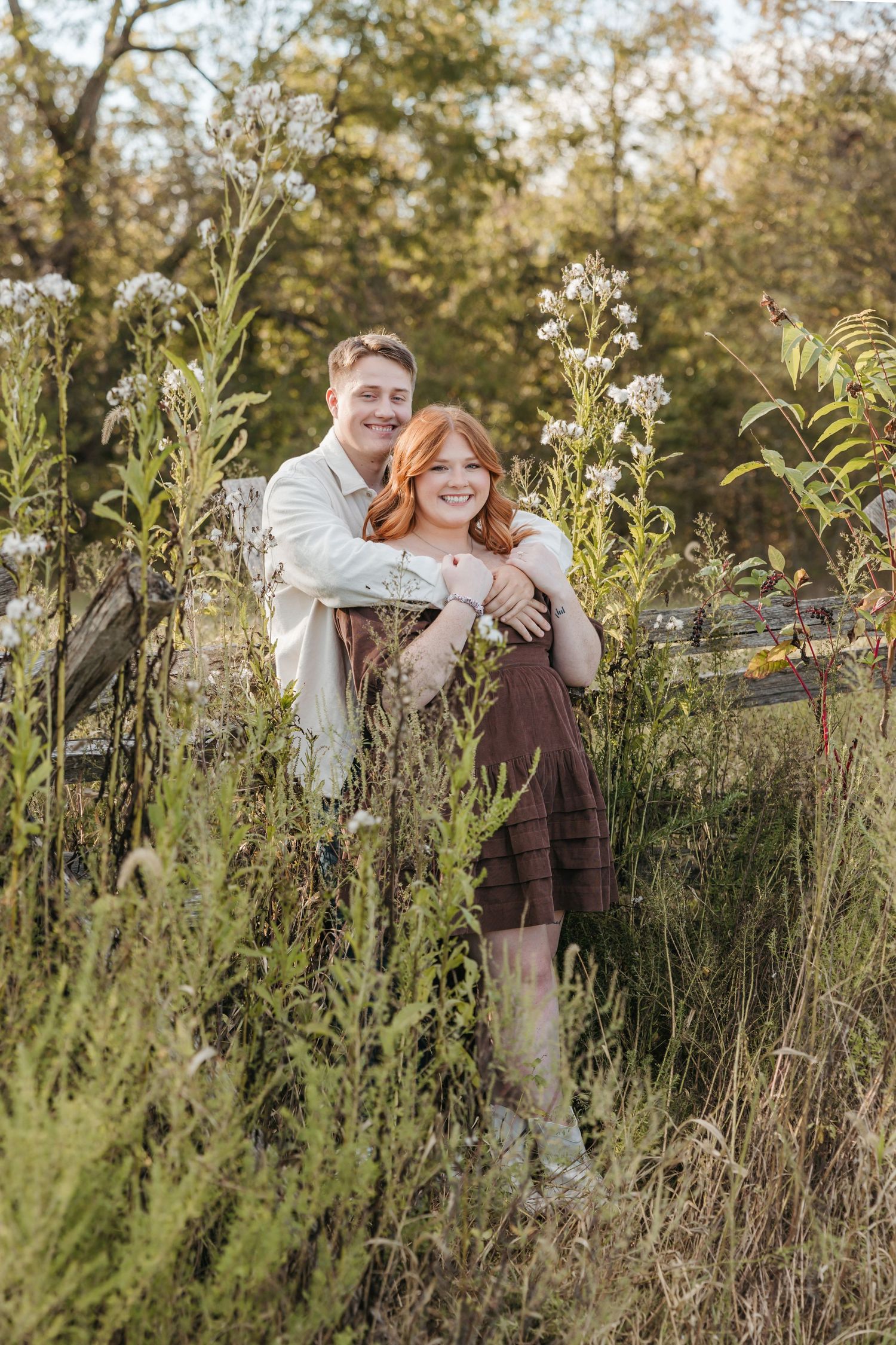 A couple embraces in a sunlit field surrounded by tall wildflowers and natural foliage.