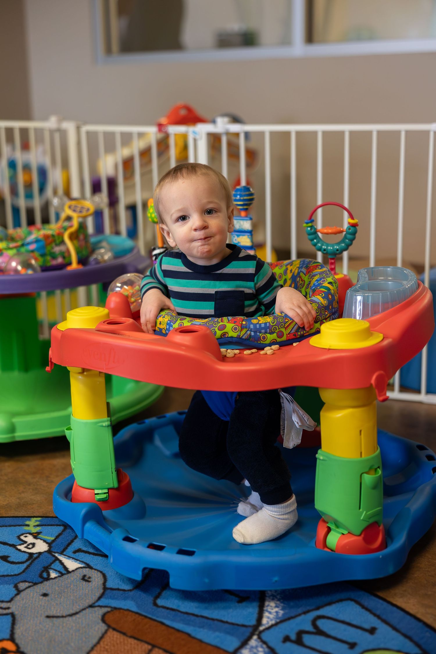 Baby playing in colorful activity center with toys on blue patterned carpet.