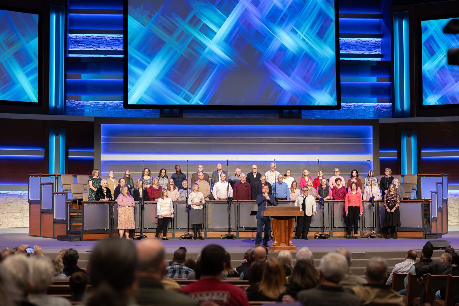 Choir group performing on stage with modern digital screens displaying blue patterns.