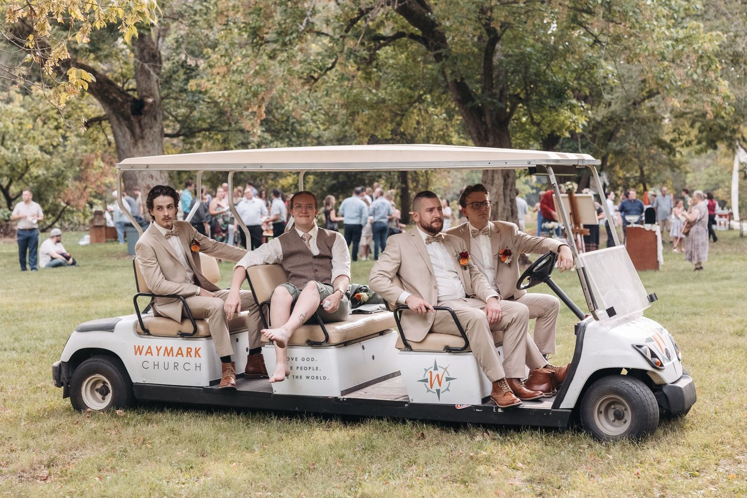 Wedding party riding in white golf cart through park setting.
