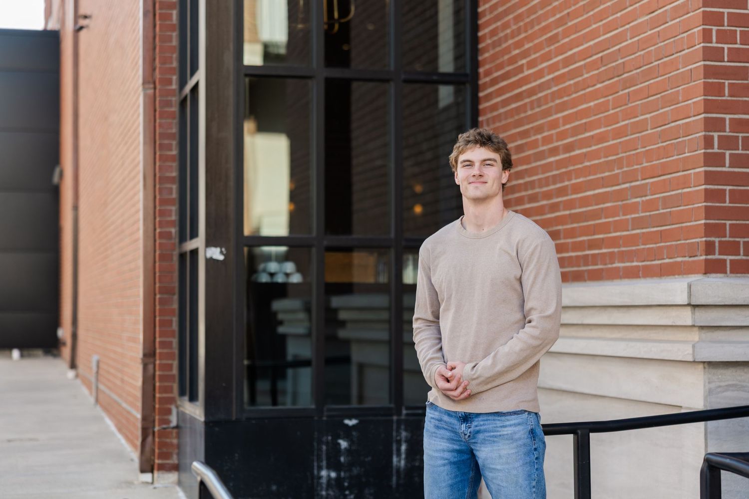 Someone wearing a light sweater and jeans stands casually in front of a brick building entrance.