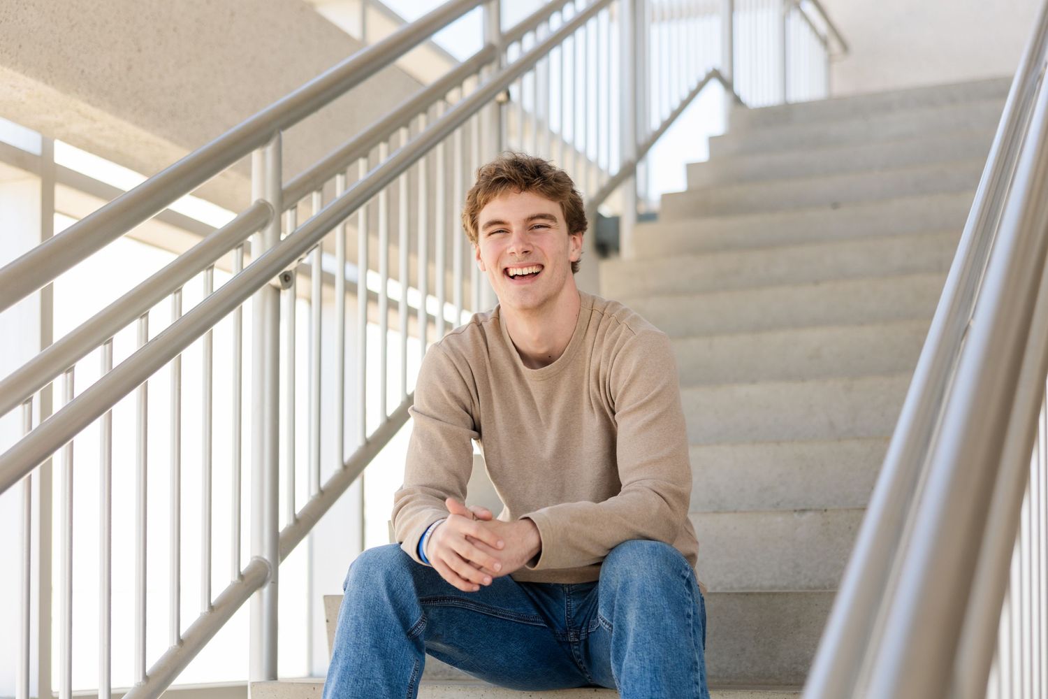 Person sits cheerfully on white staircase wearing casual beige sweater and jeans.