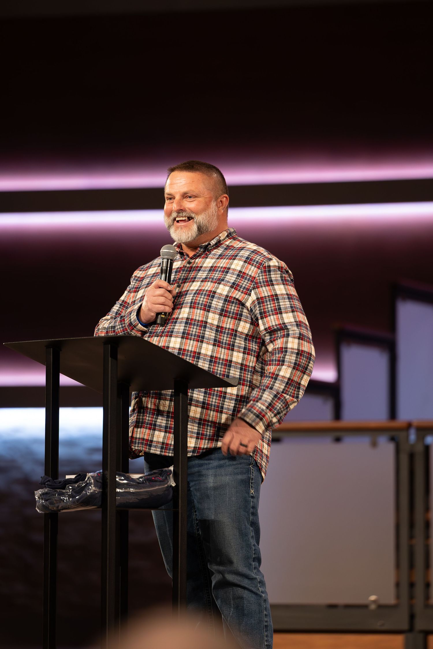 A speaker wearing a plaid flannel shirt and jeans stands at a podium on stage with pink lighting in the background.