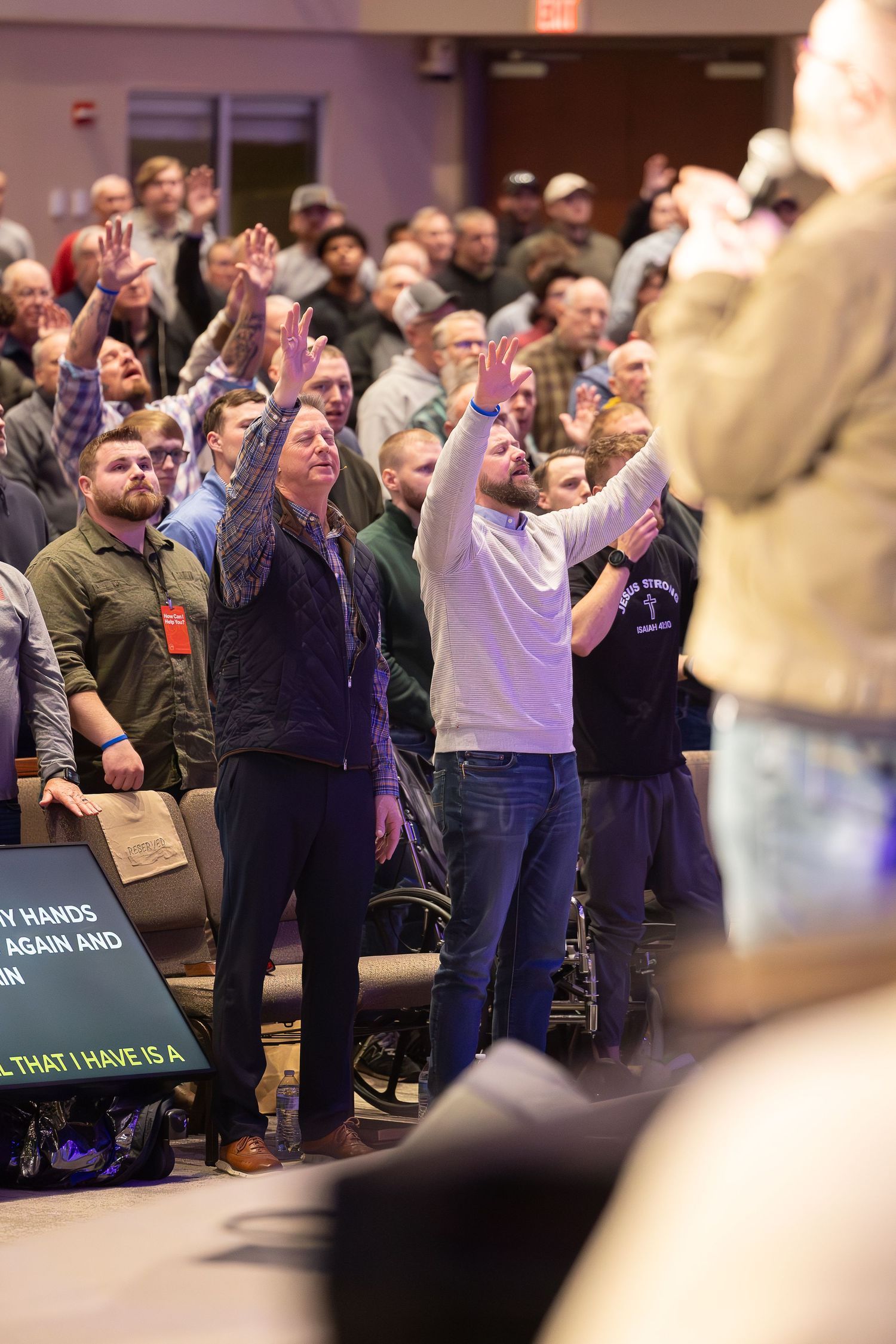 Audience members stand with raised arms during a live performance in a crowded indoor venue.