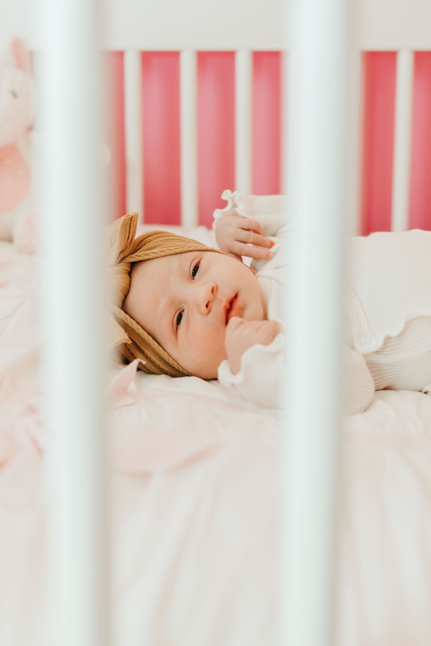 Baby sleeping peacefully in a white crib with pink walls in the background.