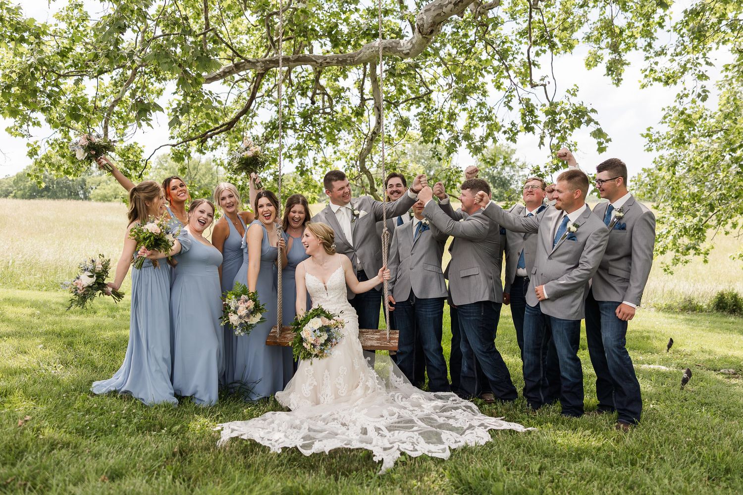 A wedding party in light blue dresses and gray suits poses together under a blooming tree in a sunny field.