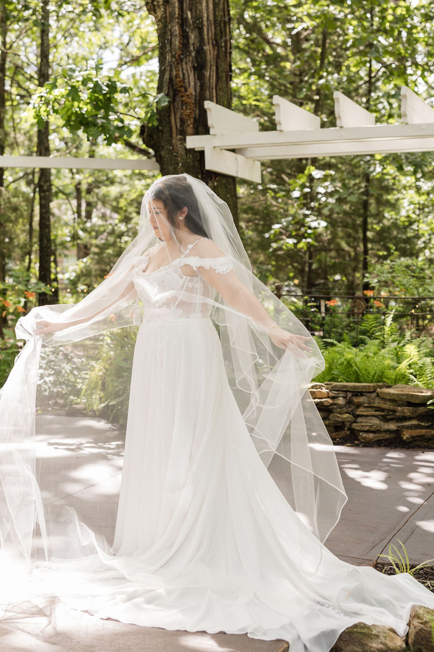 A graceful figure in a flowing white wedding gown and veil poses outdoors in a sunlit wooded garden setting.