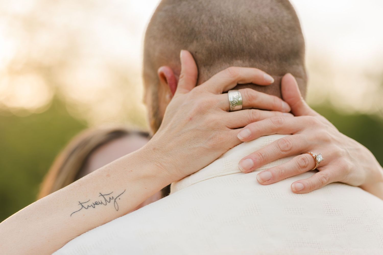 Close up view of hands embracing neck with visible script tattoo and engagement ring in warm outdoor lighting.