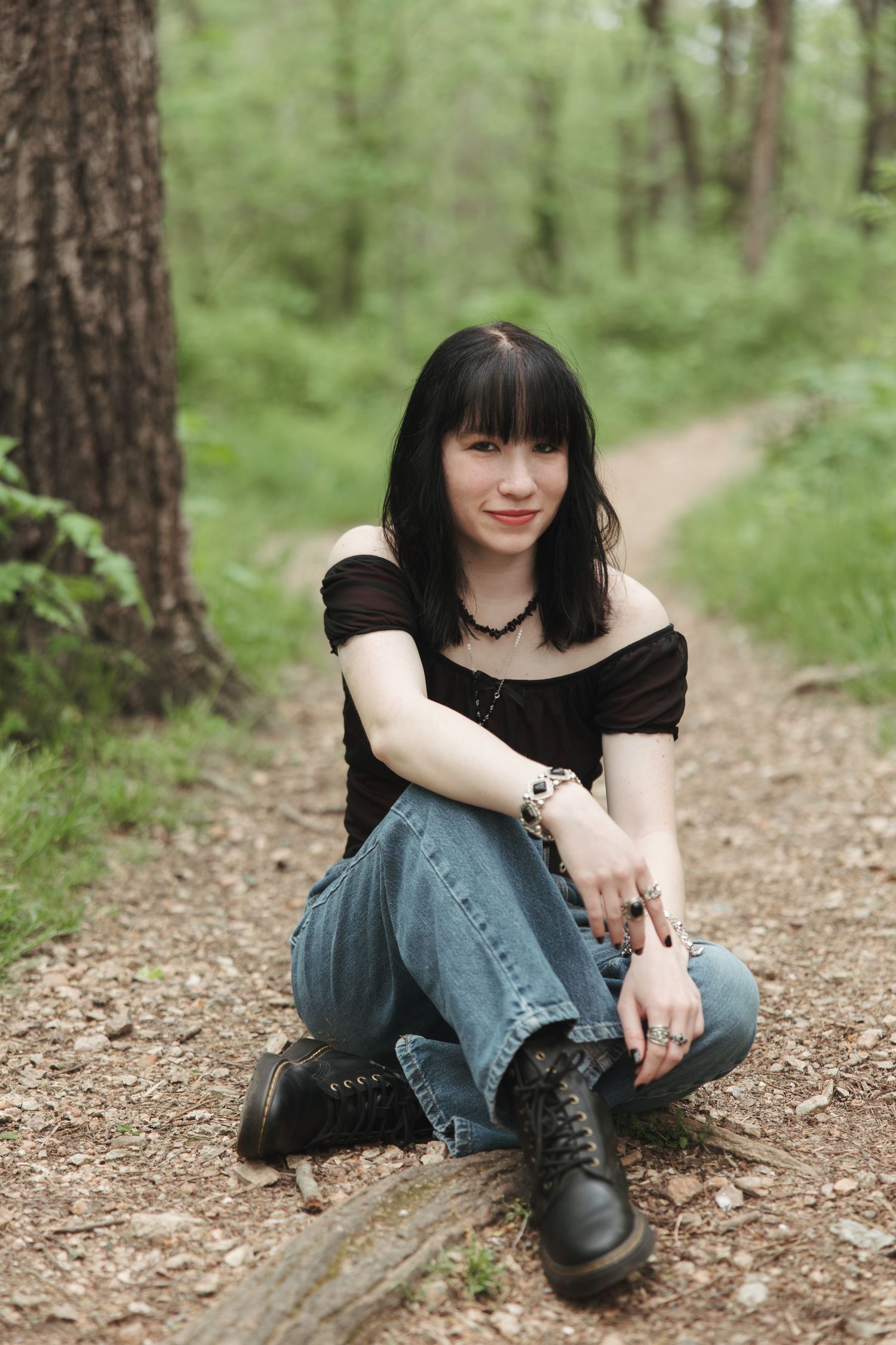 A person in a black top and jeans sits casually on a forest trail wearing combat boots.